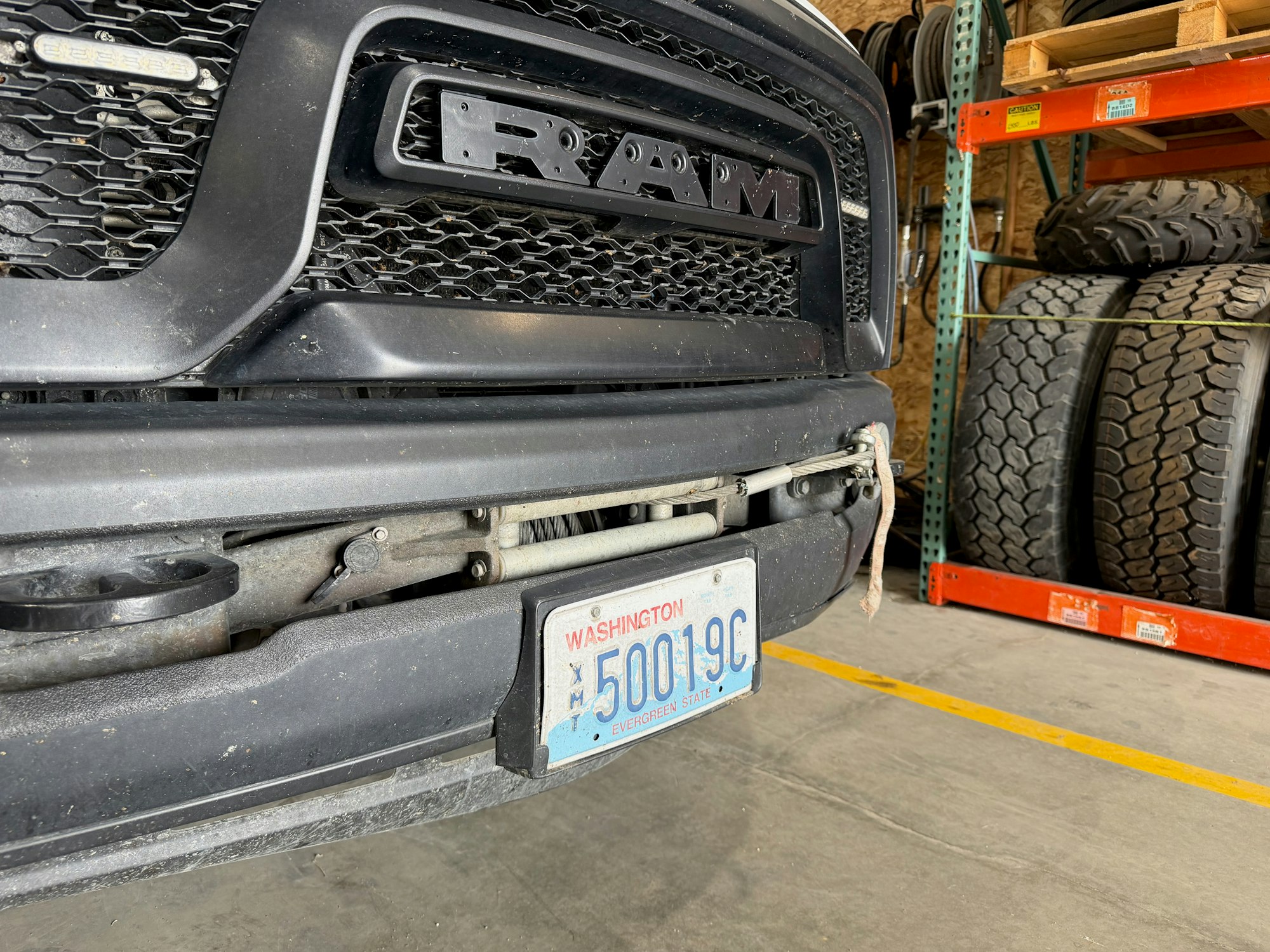 A close-up of a RAM truck's front end, showing the bumper, license plate from Washington, and shelves with tires in the background.