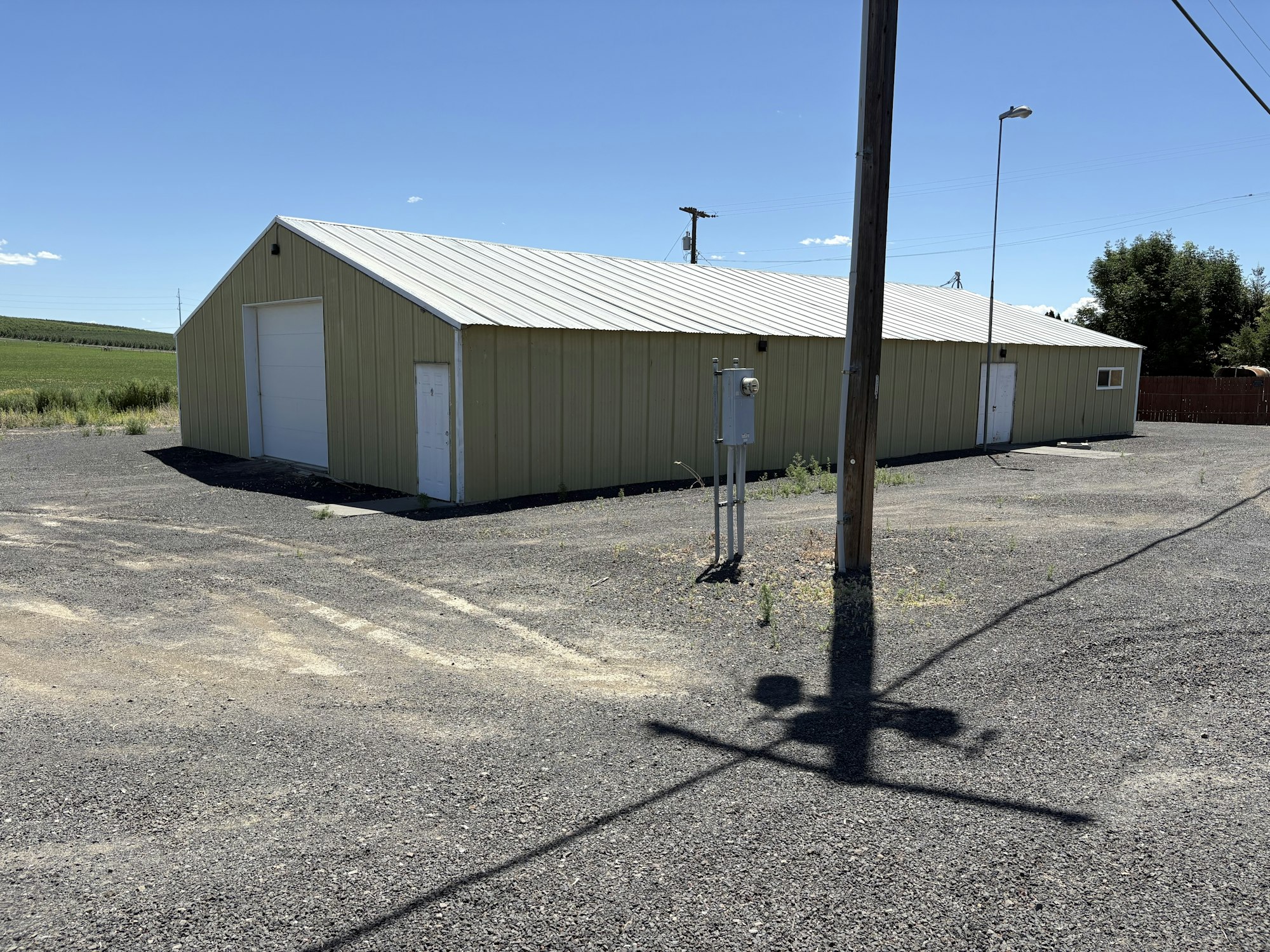 A large metal-paneled building with a garage door, surrounded by a gravel lot, utility poles, and open fields.