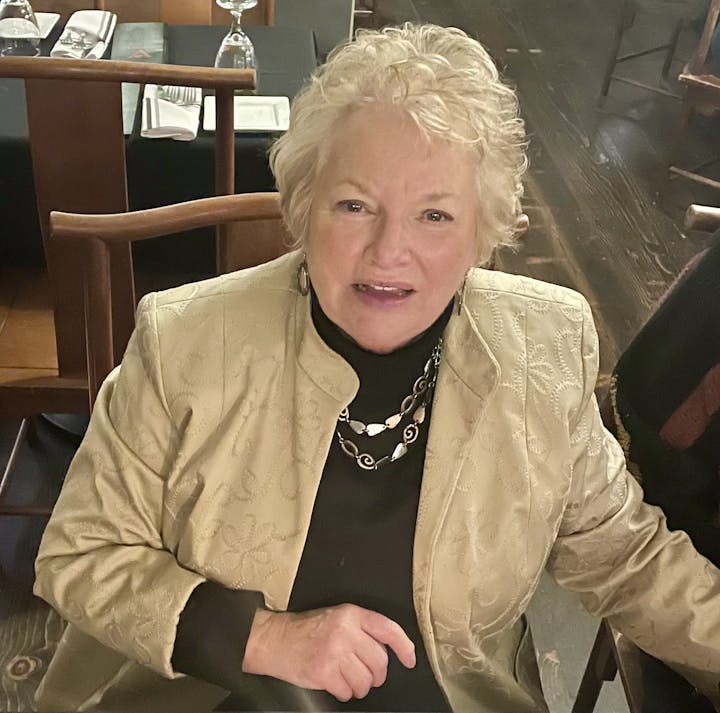 A person with short, light-colored hair sits at a table in a restaurant, wearing a patterned jacket and a necklace.
