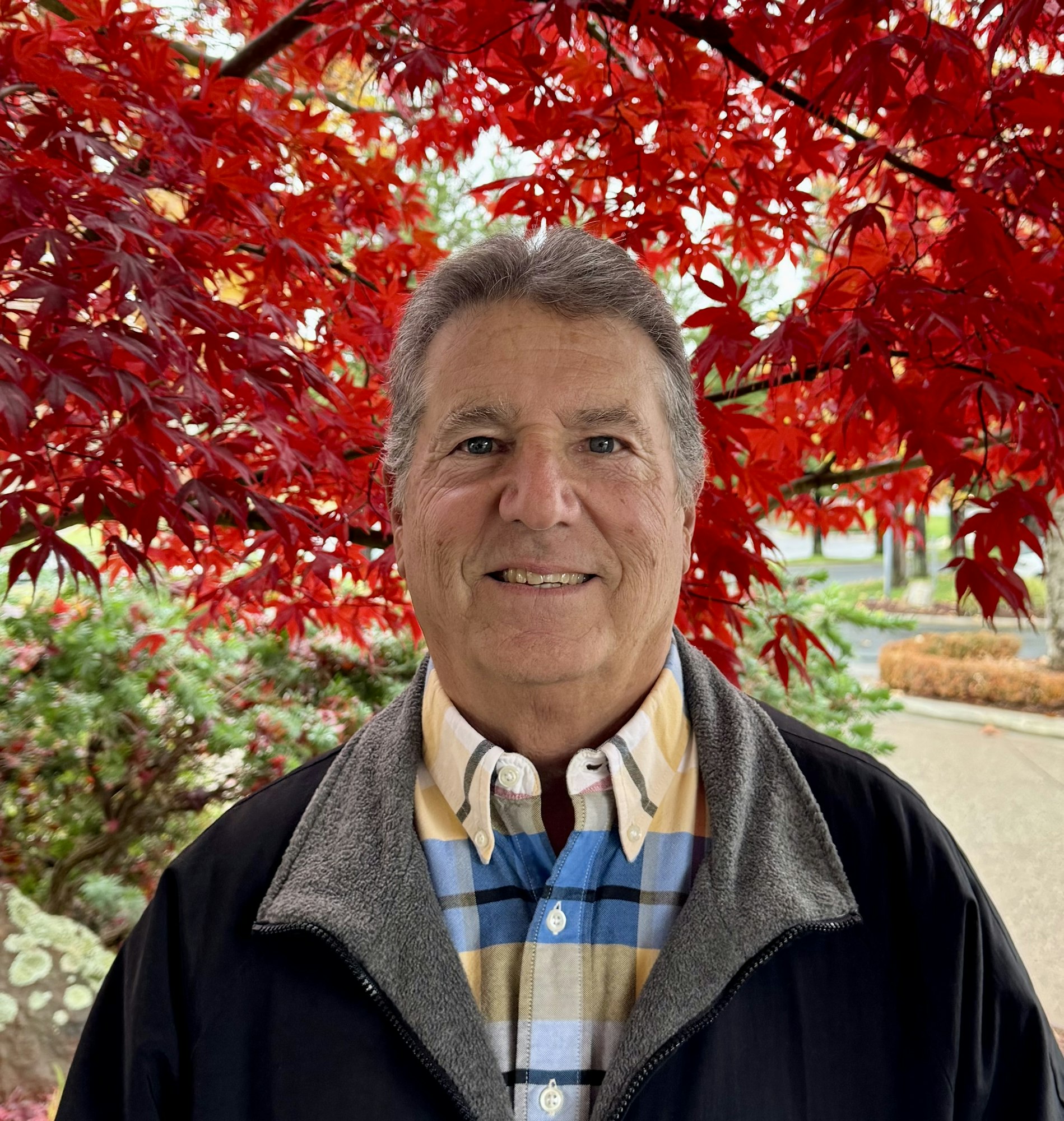 A smiling man stands in front of bright red autumn leaves.