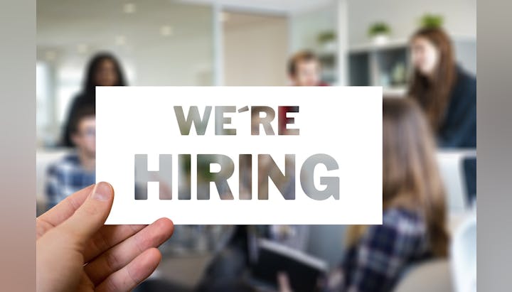 A sign with the words "WE'RE HIRING" held in front of people in an office.