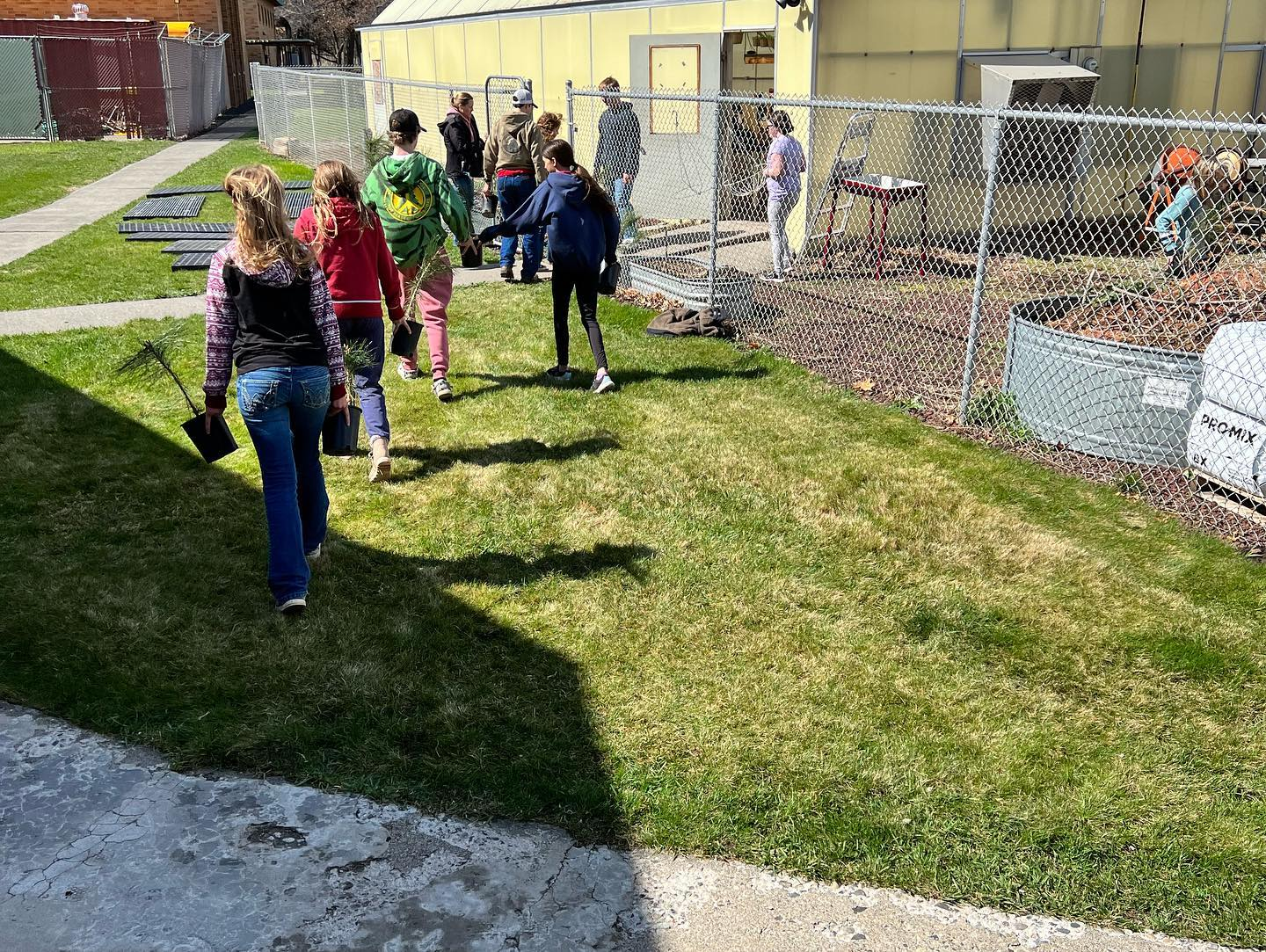 Group of people walking outside near a chain-link fence, some holding tools, possibly for a gardening or outdoor activity.