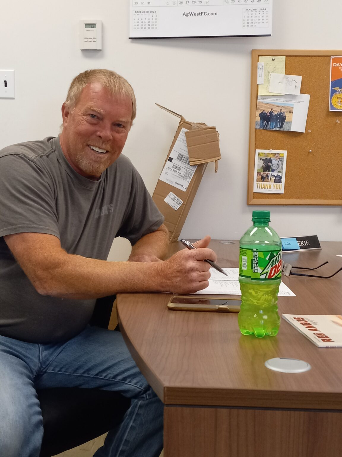 A smiling man sitting at a desk, writing on a paper, with a bottle of Mountain Dew in the foreground.