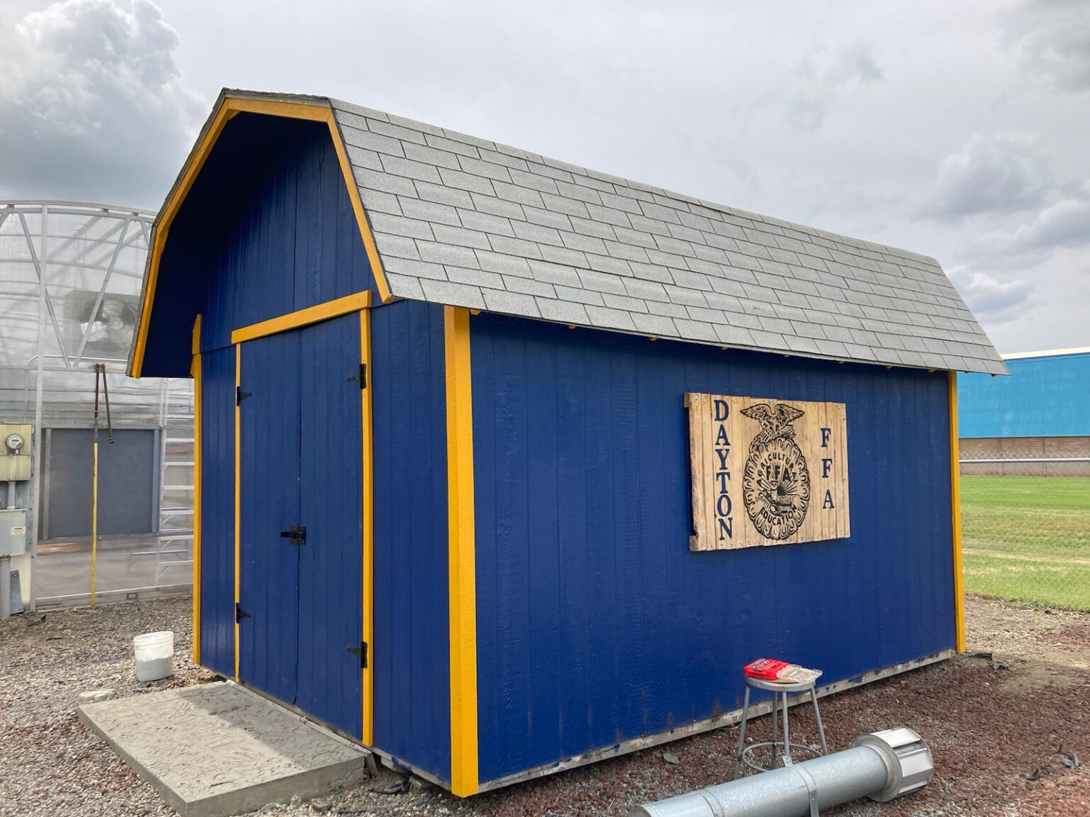 A blue shed with a wooden FFA emblem, yellow trim, and contrasting grey roof.