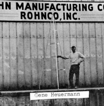 A person stands by a ladder in front of a sign for John Manufacturing Co., Rohnco, Inc., with "Gene Heuermann" captioned below.
