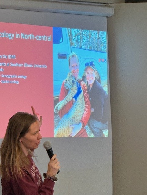 A person is giving a presentation with a slide showing two others holding an animal, with text about ecology.