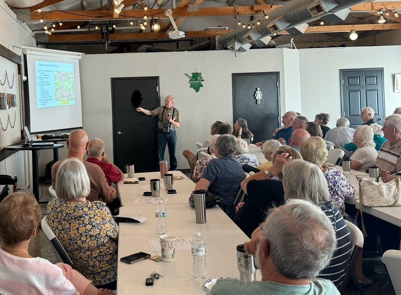 Lecture on birding with an audience and a presenter pointing at a screen in a room with industrial-style lighting.