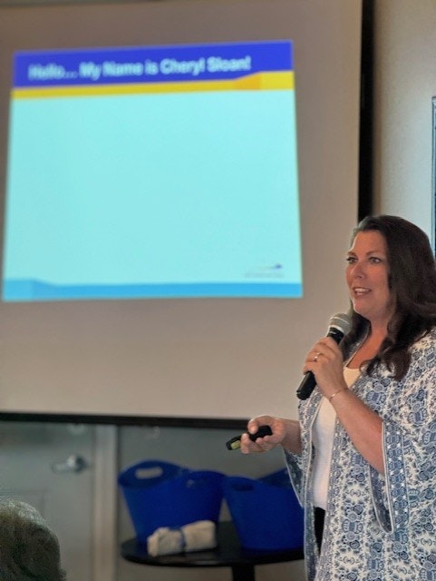 A woman is presenting with a microphone, standing in front of a projector screen that reads "Hello...My Name is Cheryl Steck".