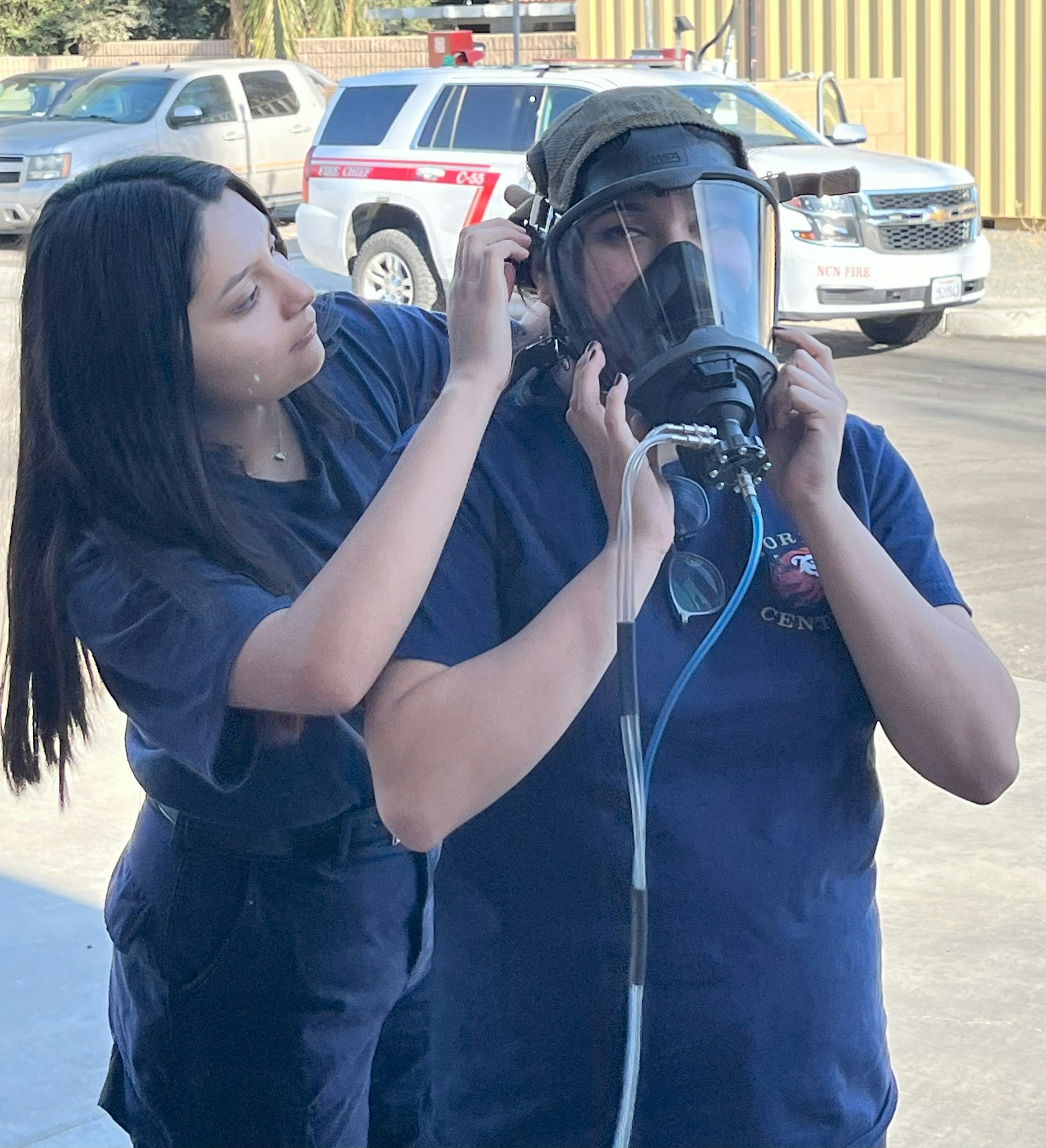 Two people in blue uniforms, one adjusting a gas mask on the other, in front of emergency vehicles.