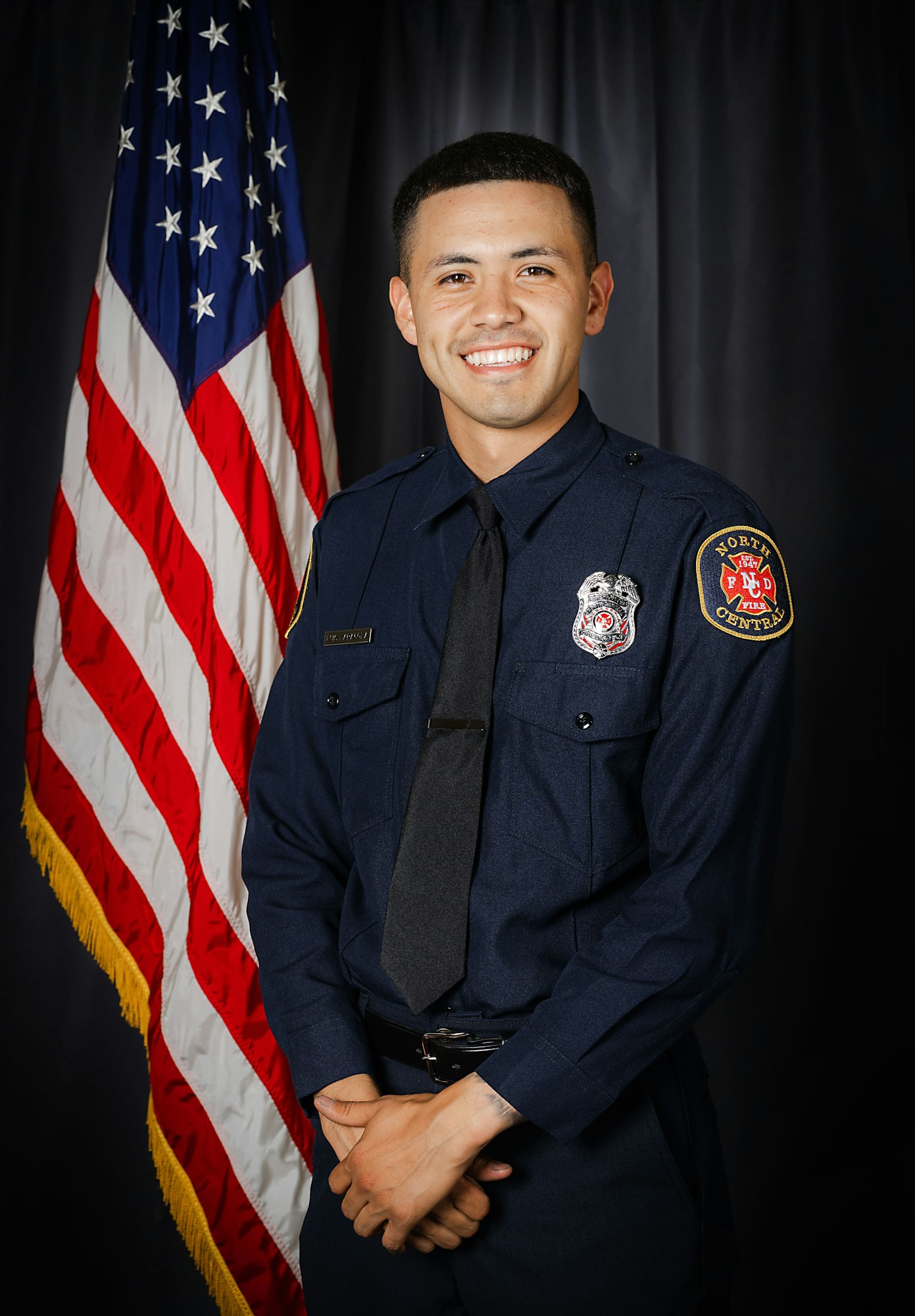A smiling person in a firefighter's uniform standing in front of an American flag.