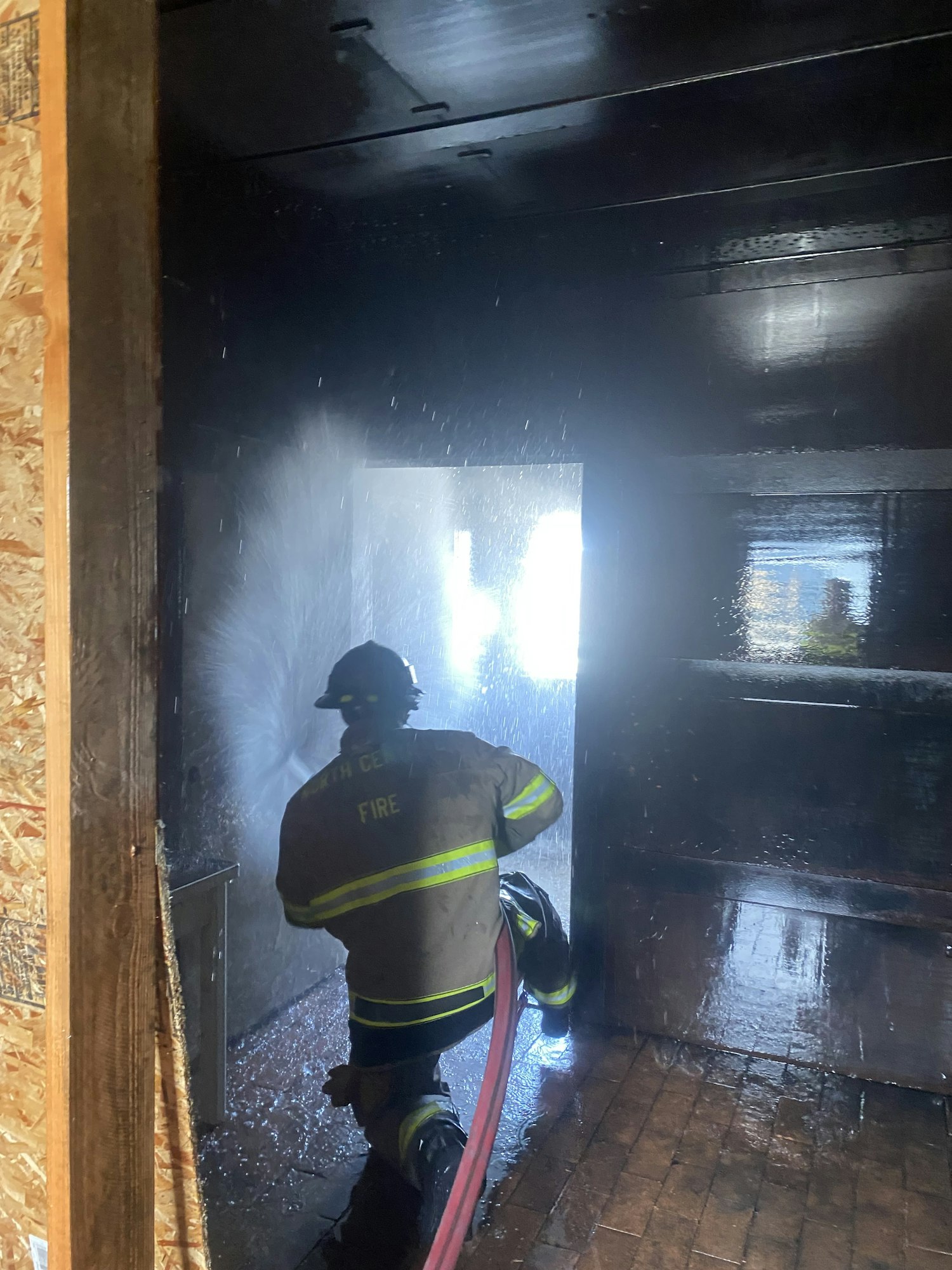 A firefighter is battling a fire inside a dark room, spraying water while water droplets are visible in the air.
