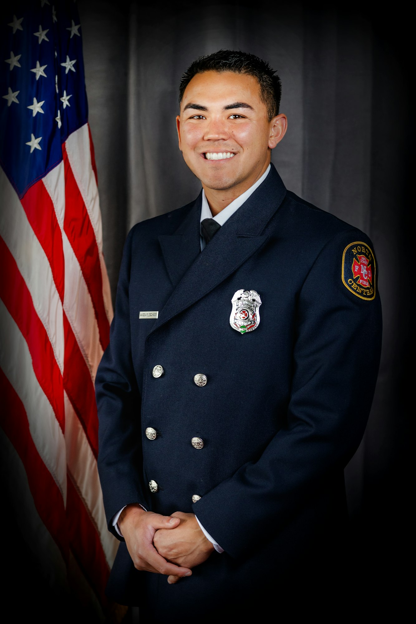 A person in a formal uniform stands beside an American flag, smiling.