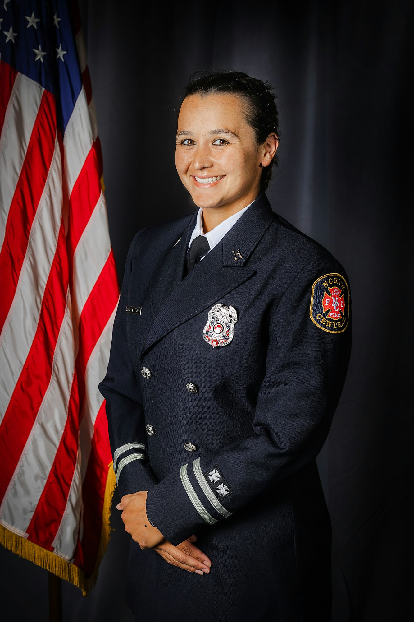 A person in a firefighter dress uniform stands beside an American flag, smiling.
