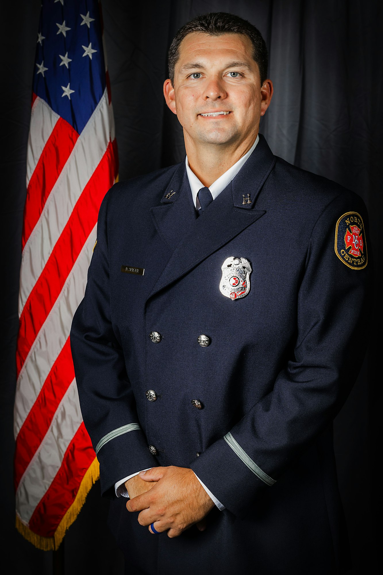 A person in a dark uniform with a badge stands in front of an American flag, hands clasped.