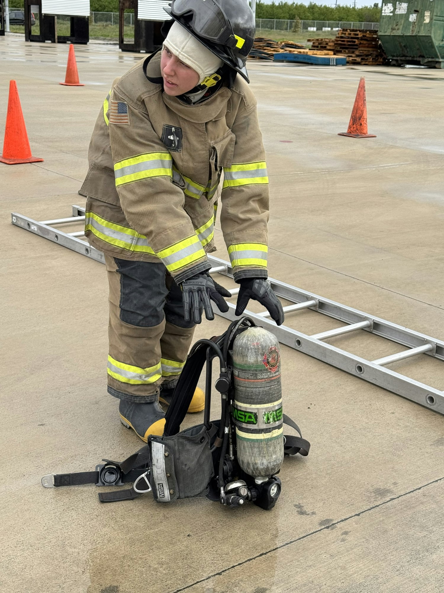 A firefighter is kneeling beside a ladder and an air tank on the ground, surrounded by traffic cones.