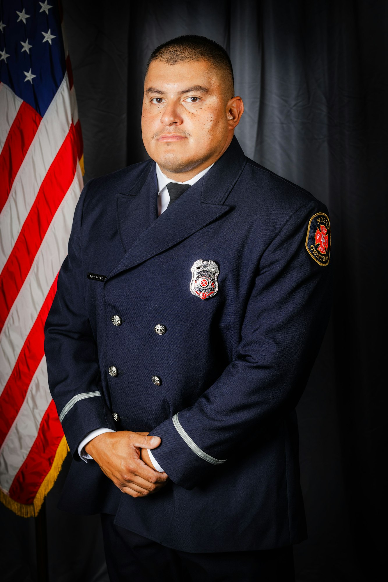 A person in a formal uniform stands by an American flag, against a dark backdrop.