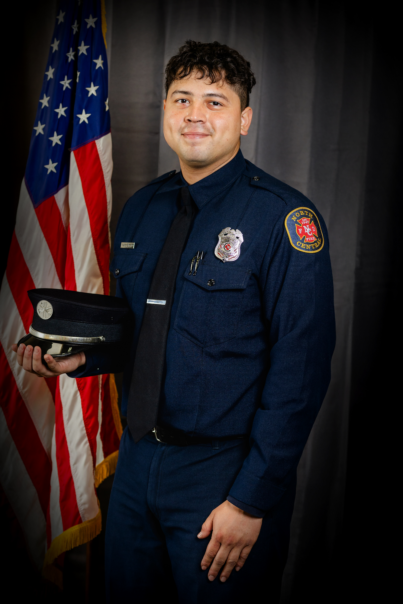 Fabio Quijano in a dark uniform stands beside an American flag, against a dark curtain background.