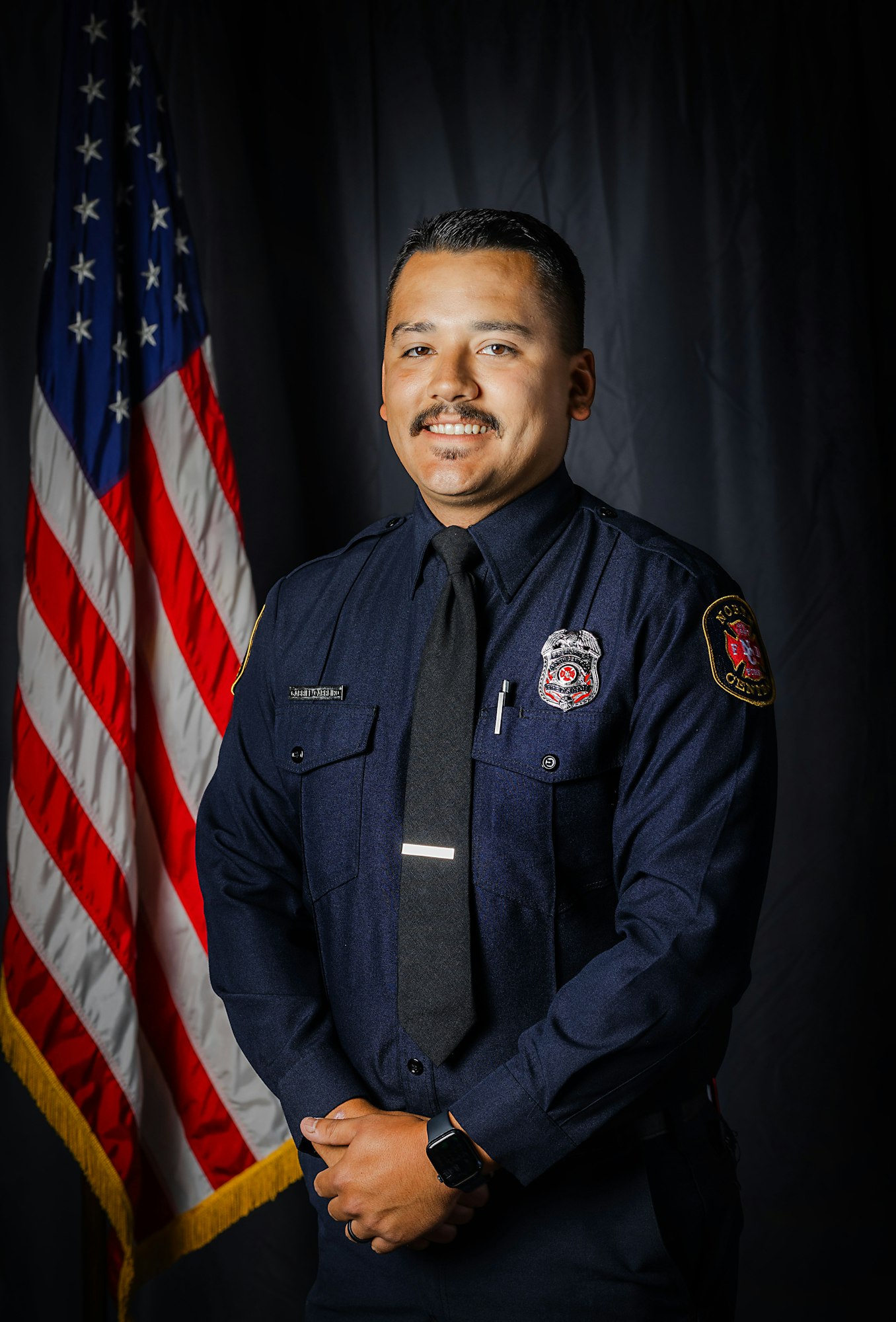 A smiling firefighter in a dark uniform stands in front of the American flag.