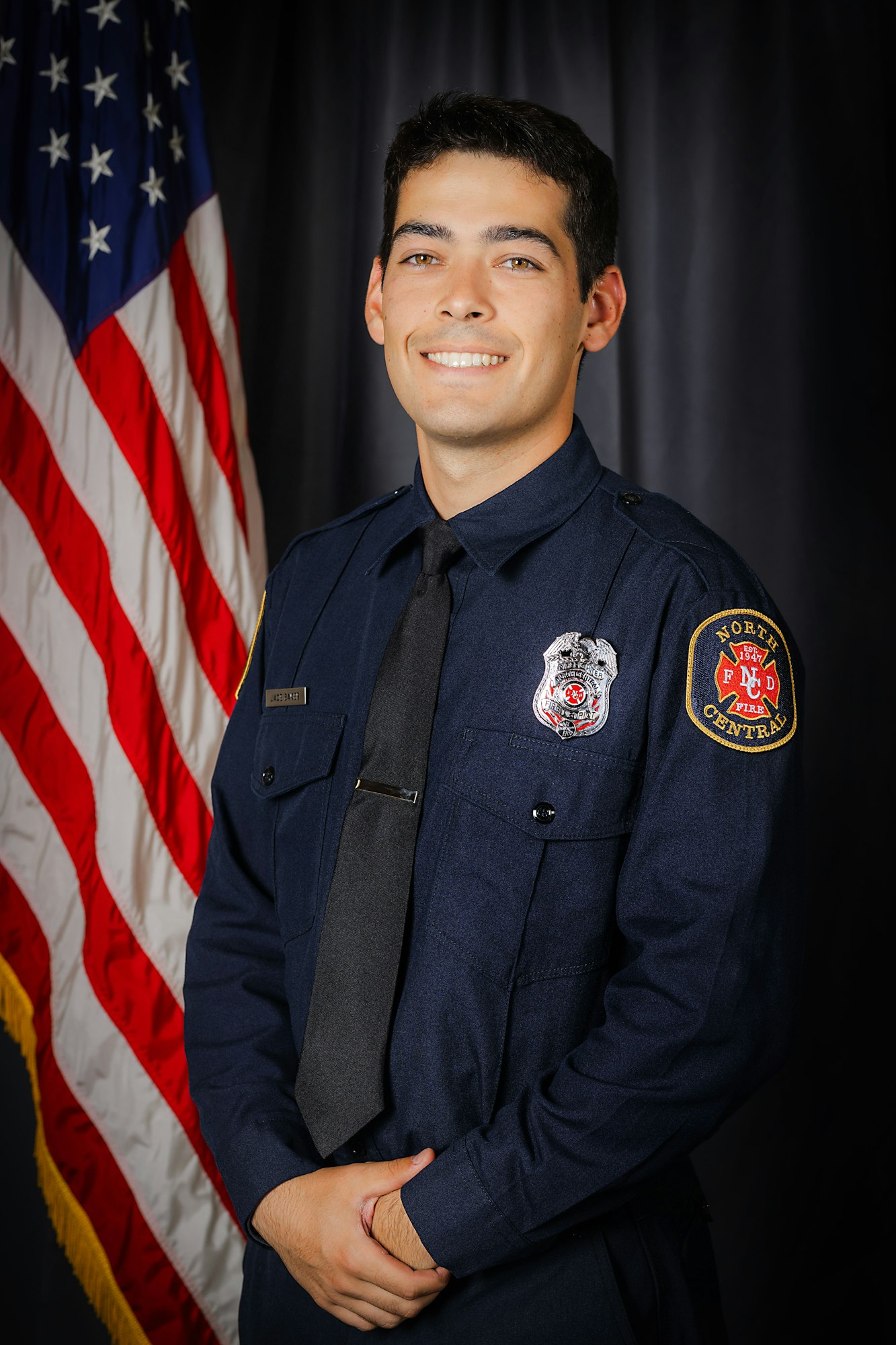 A smiling firefighter in uniform in front of an American flag.