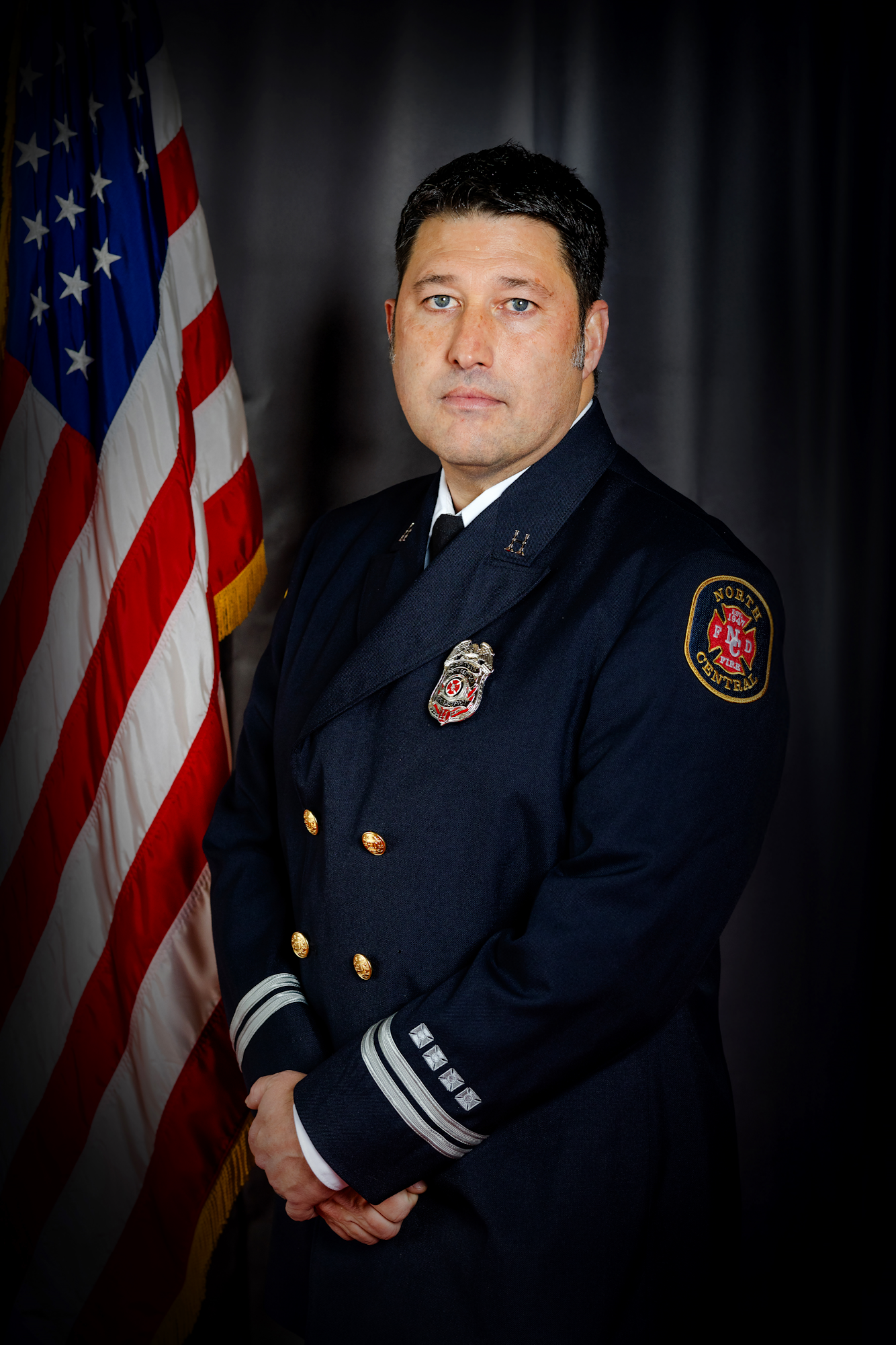 A person in a formal uniform stands by an American flag, against a dark backdrop.