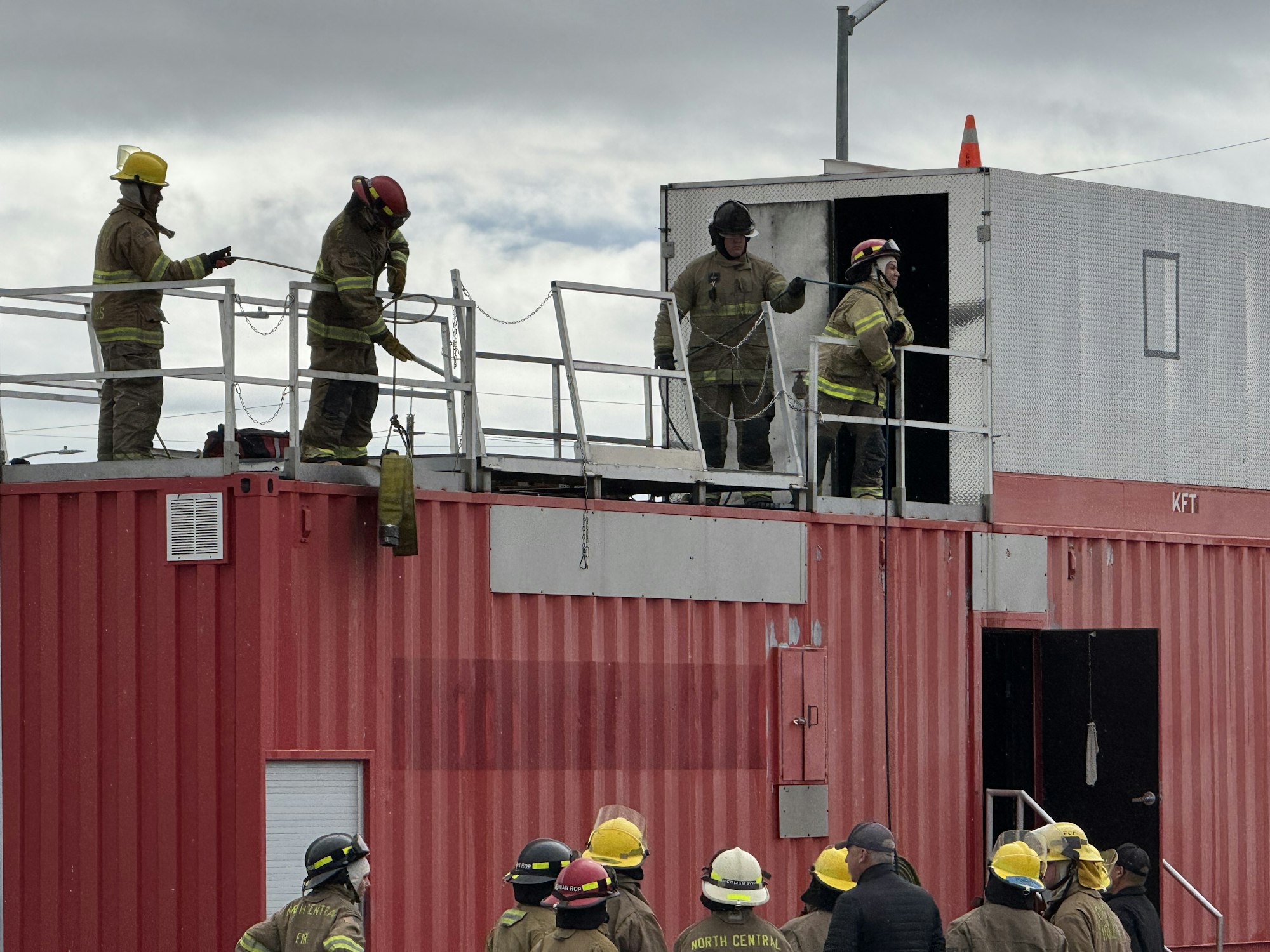 Firefighters on a training structure, some on the roof while others watch from below, wearing protective gear and helmets.