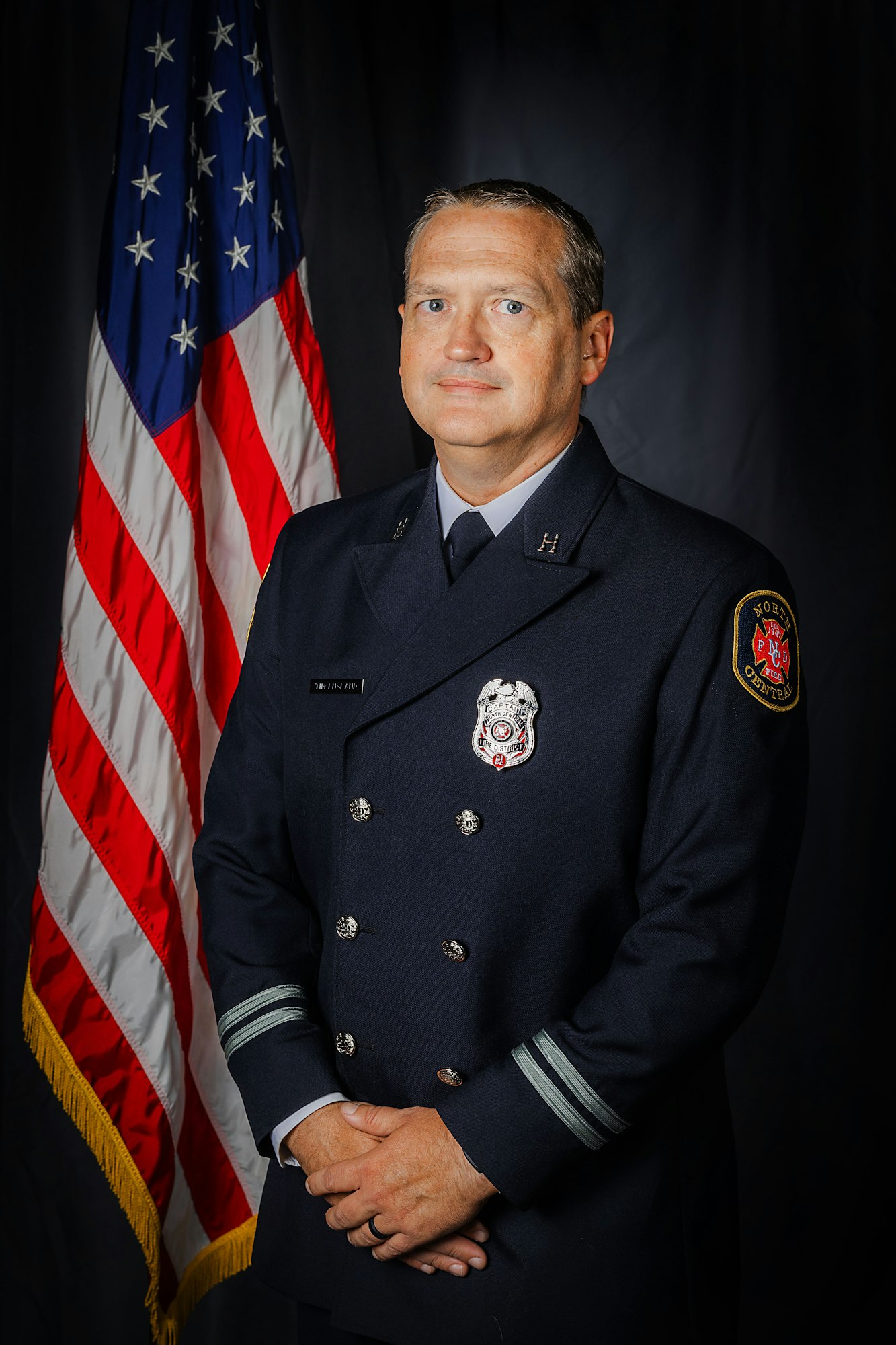 A person in a formal firefighter uniform stands in front of an American flag.