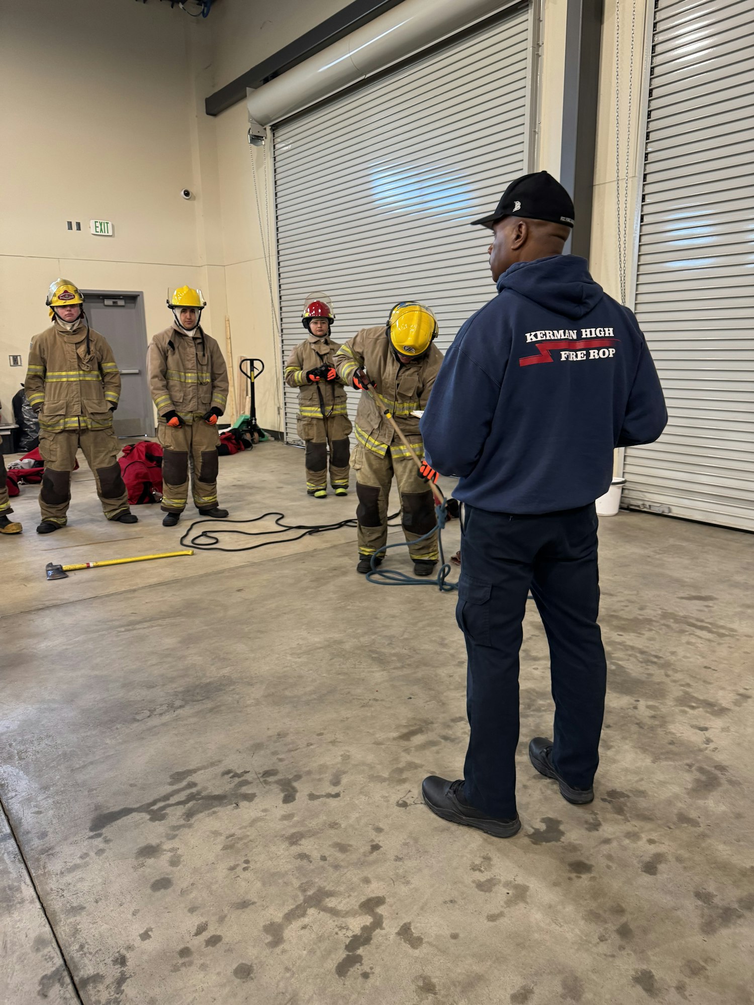 A group of people in firefighting gear being observed by a person in a sweatshirt indoors.