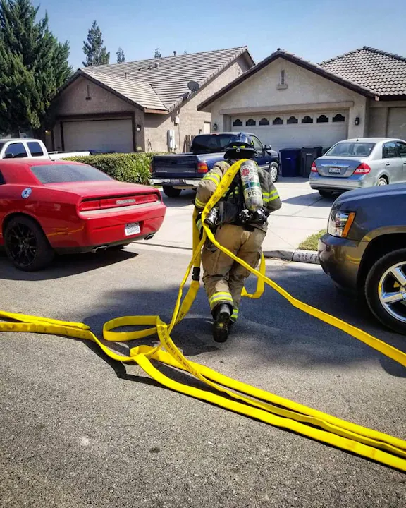 A firefighter is walking on a street, carrying yellow hoses near parked cars and residential houses.