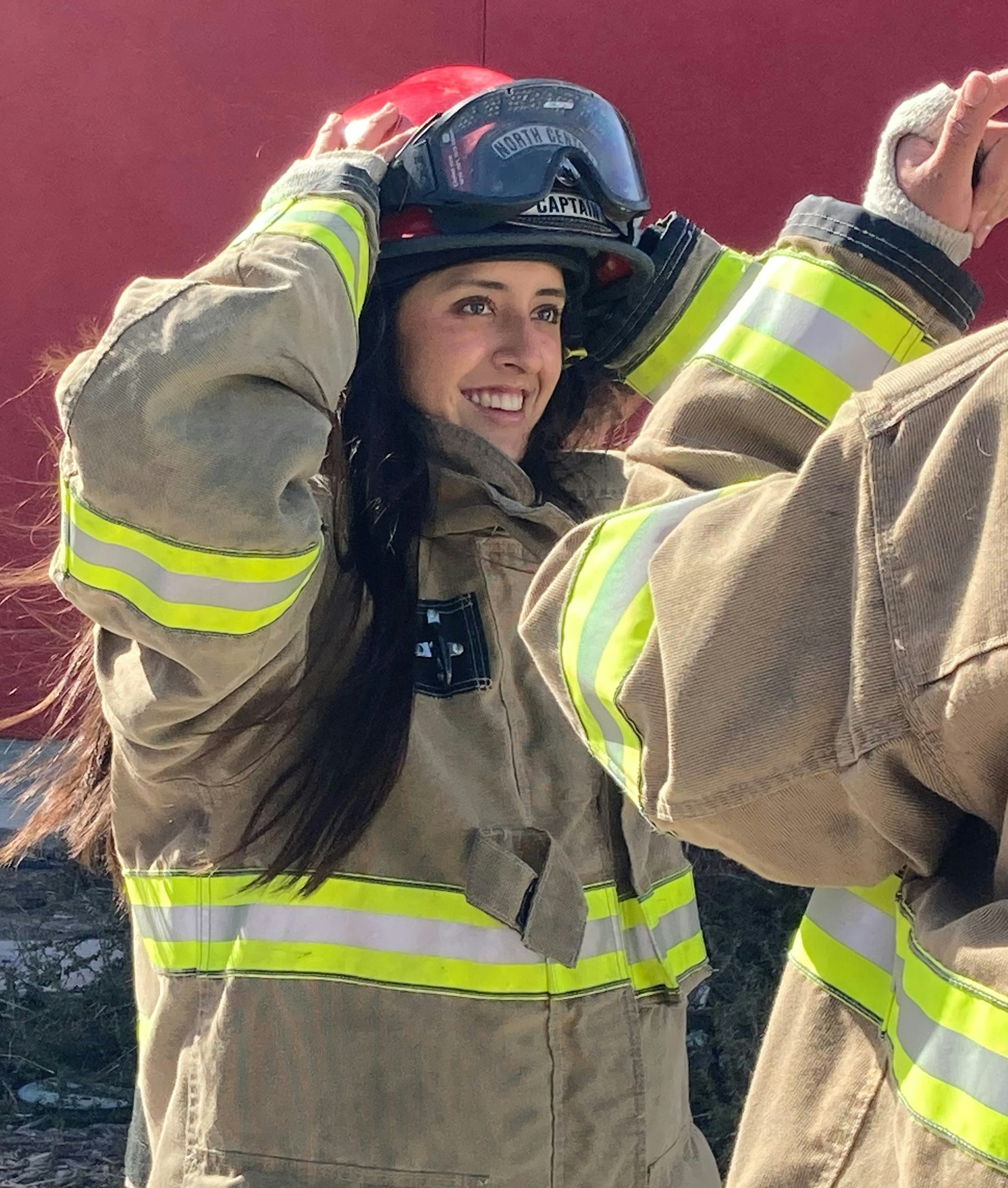 A smiling person in firefighter gear adjusts their helmet, ready for action, while another person assists nearby.
