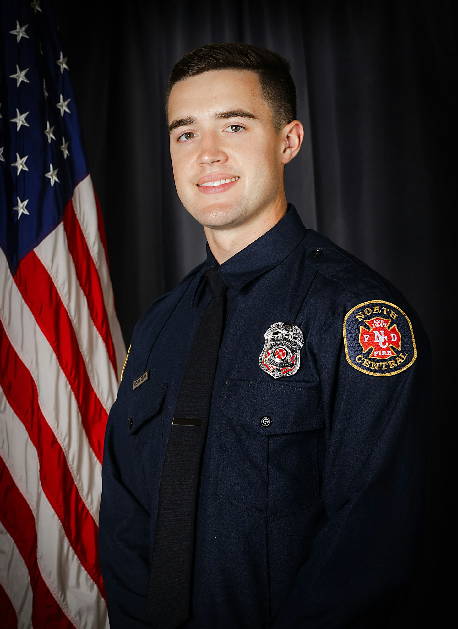 A person in a firefighter's uniform stands before an American flag.