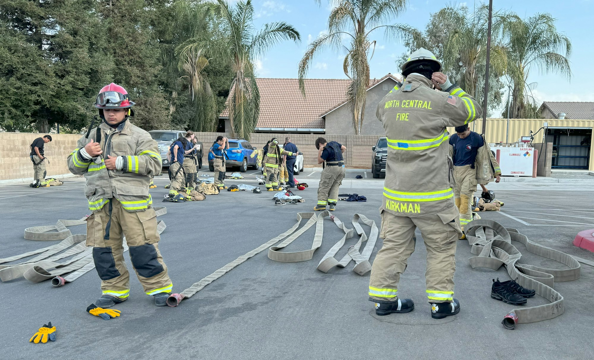 Firefighters in gear, practicing with hoses outdoors, surrounded by equipment and palm trees.