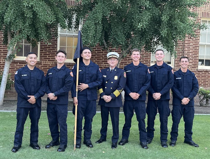 A group of seven firefighters in uniform posing for a photo outdoors, one with a flagpole, another in different attire indicating higher rank.