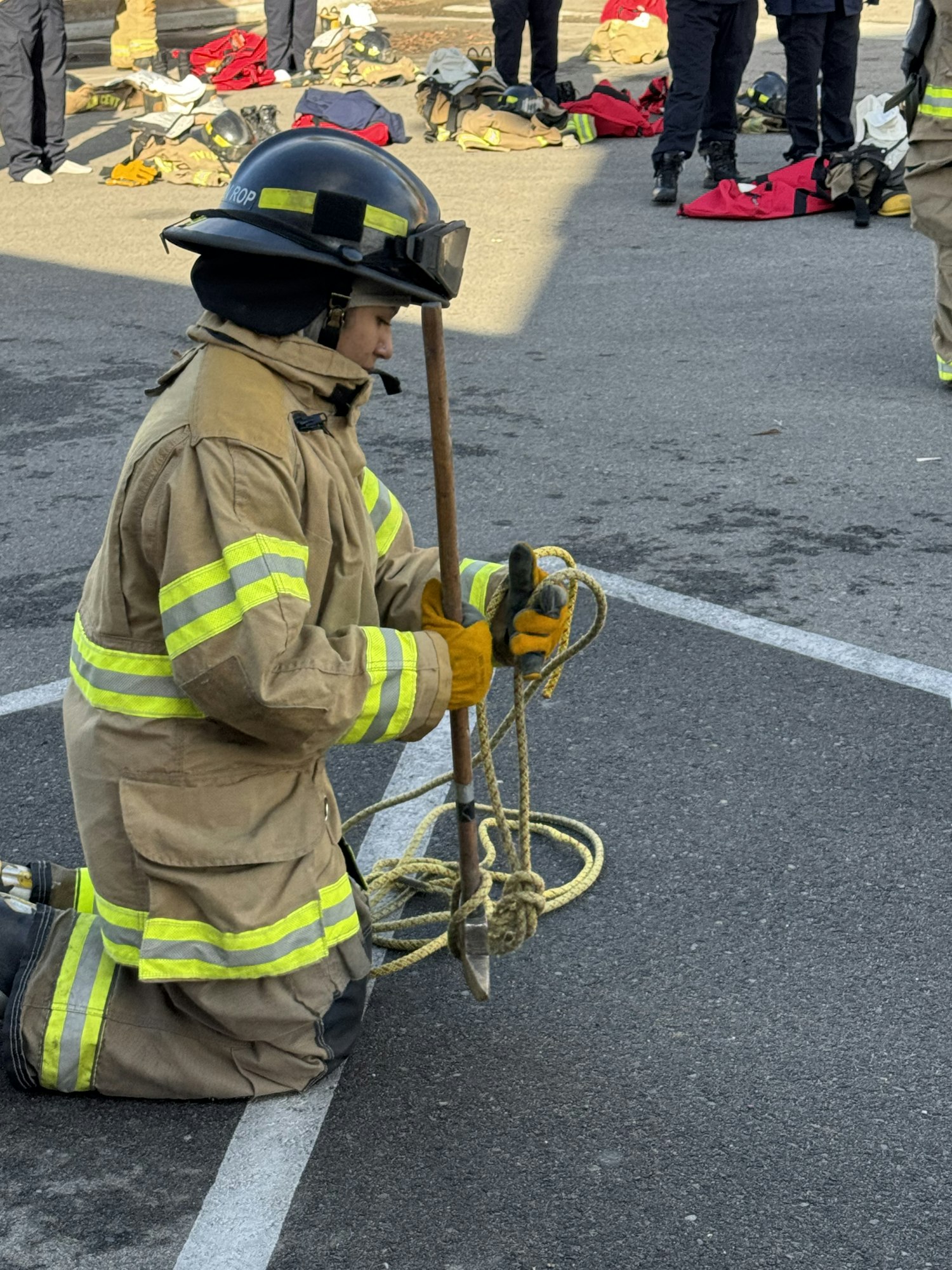 A firefighter in gear kneels on the pavement, holding a rope and tool, with equipment and people in the background.