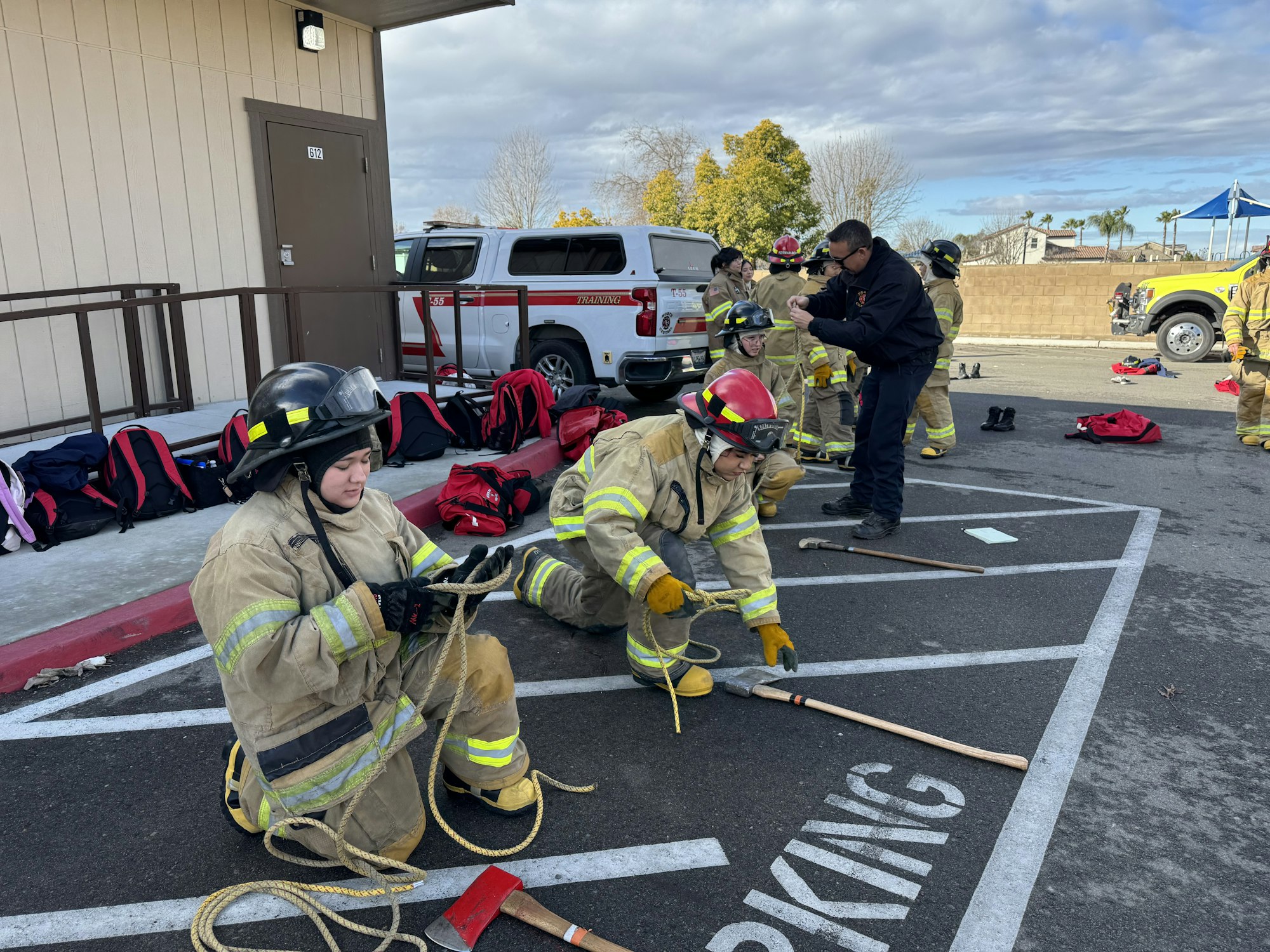 Firefighters are engaged in training outdoors, some holding ropes and tools, while others observe.