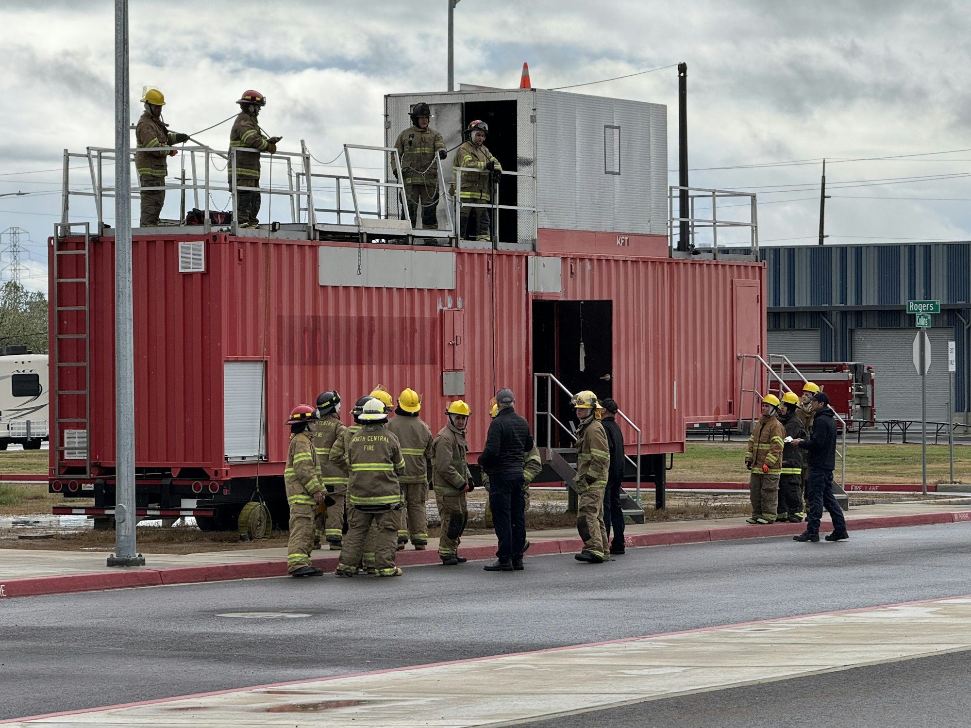 Firefighters in gear gather around a red training structure, with some on top and others on the ground.