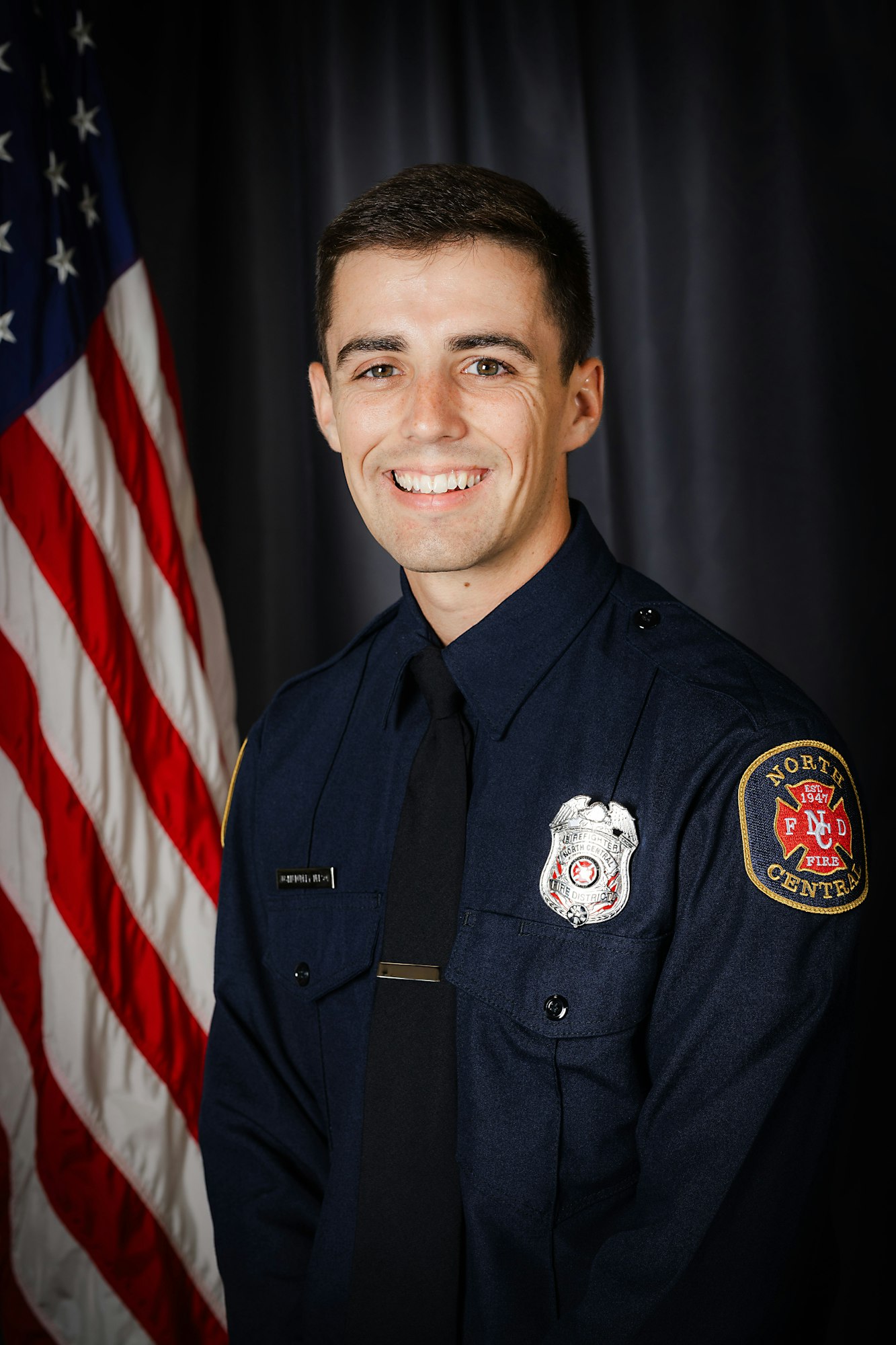 A person in a firefighter uniform with badge in front of an American flag.