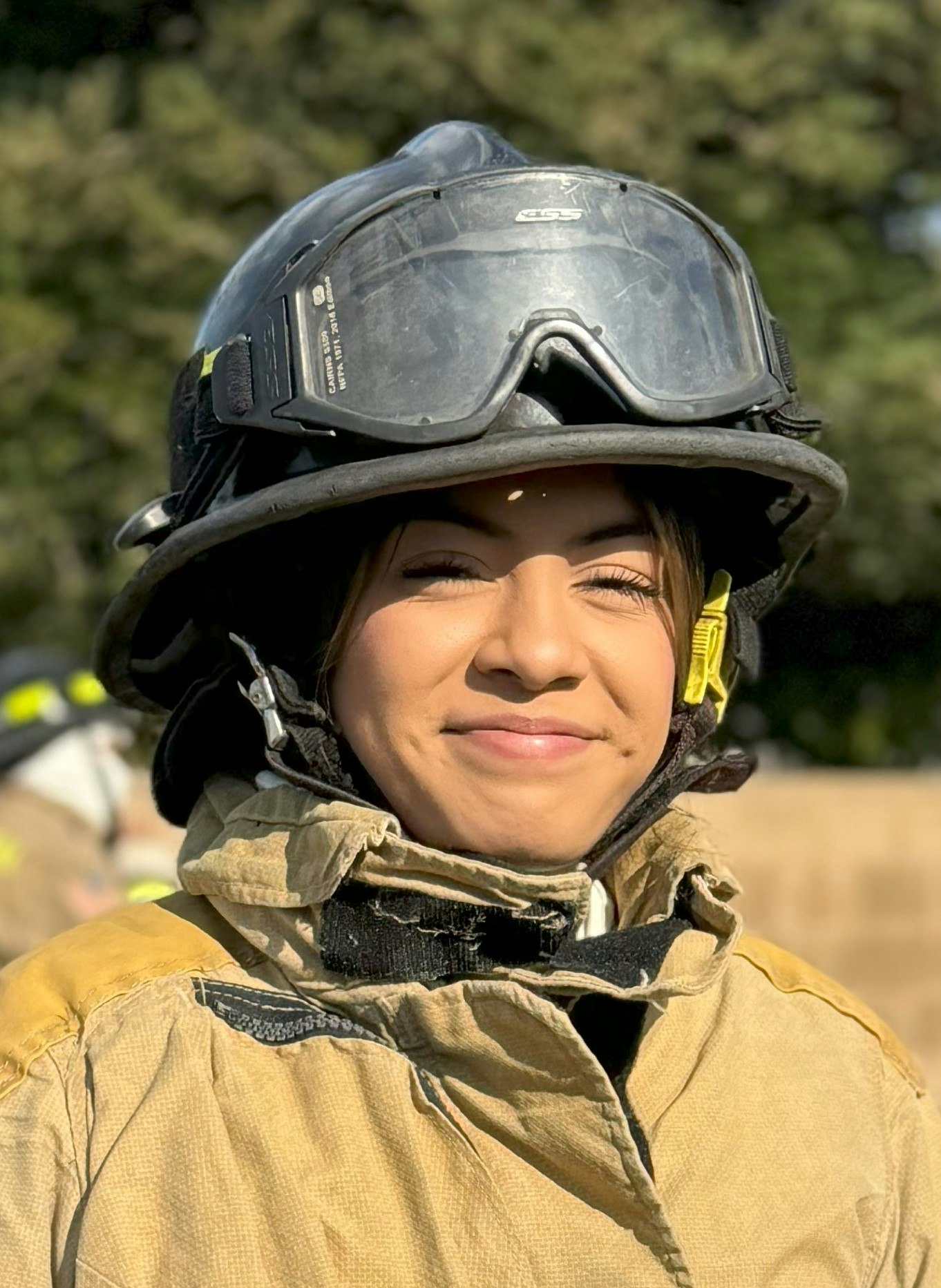 A firefighter smiling, wearing protective gear and a helmet with goggles, outdoors.