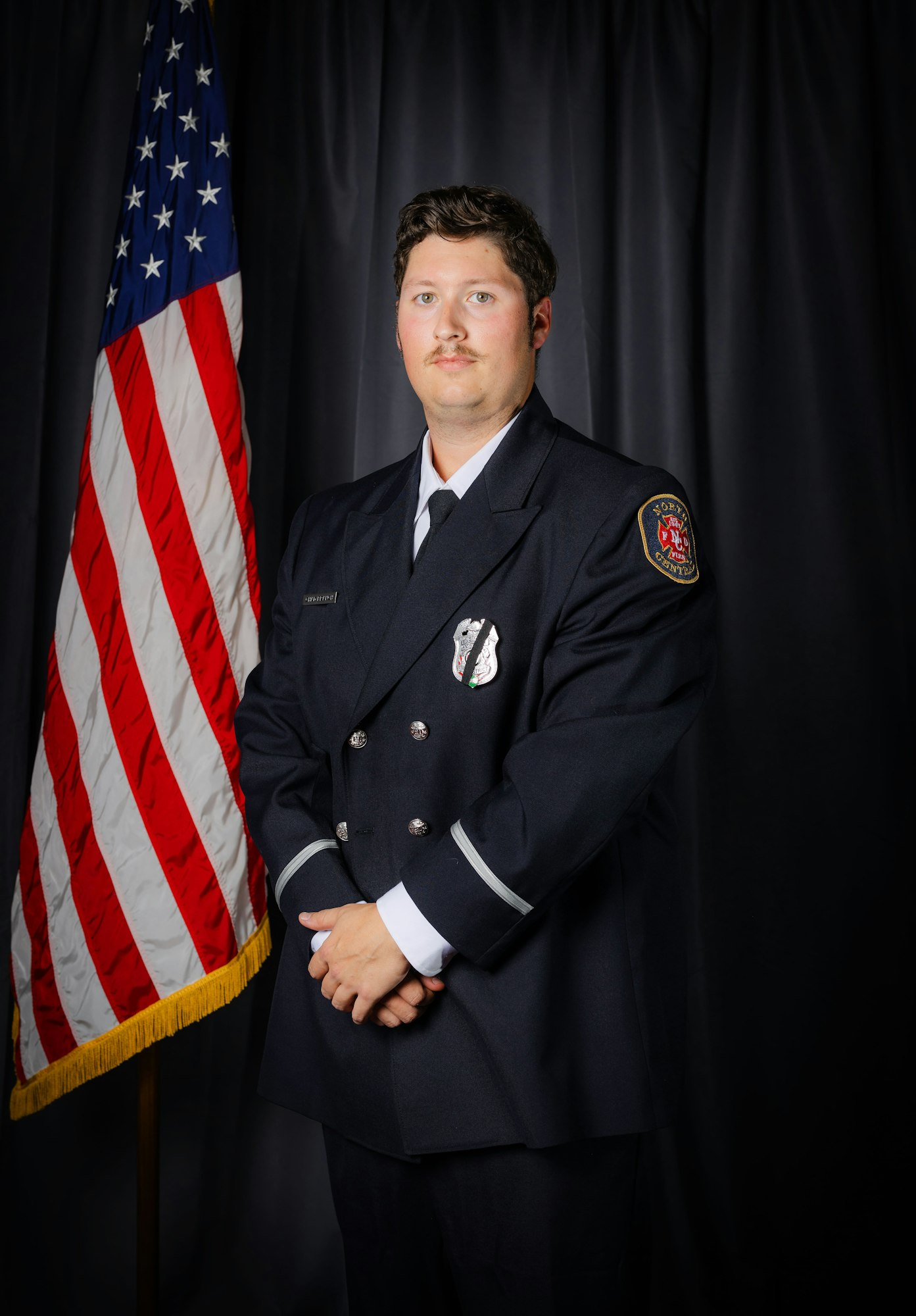 A person in a formal uniform stands beside a U.S. flag against a dark backdrop.