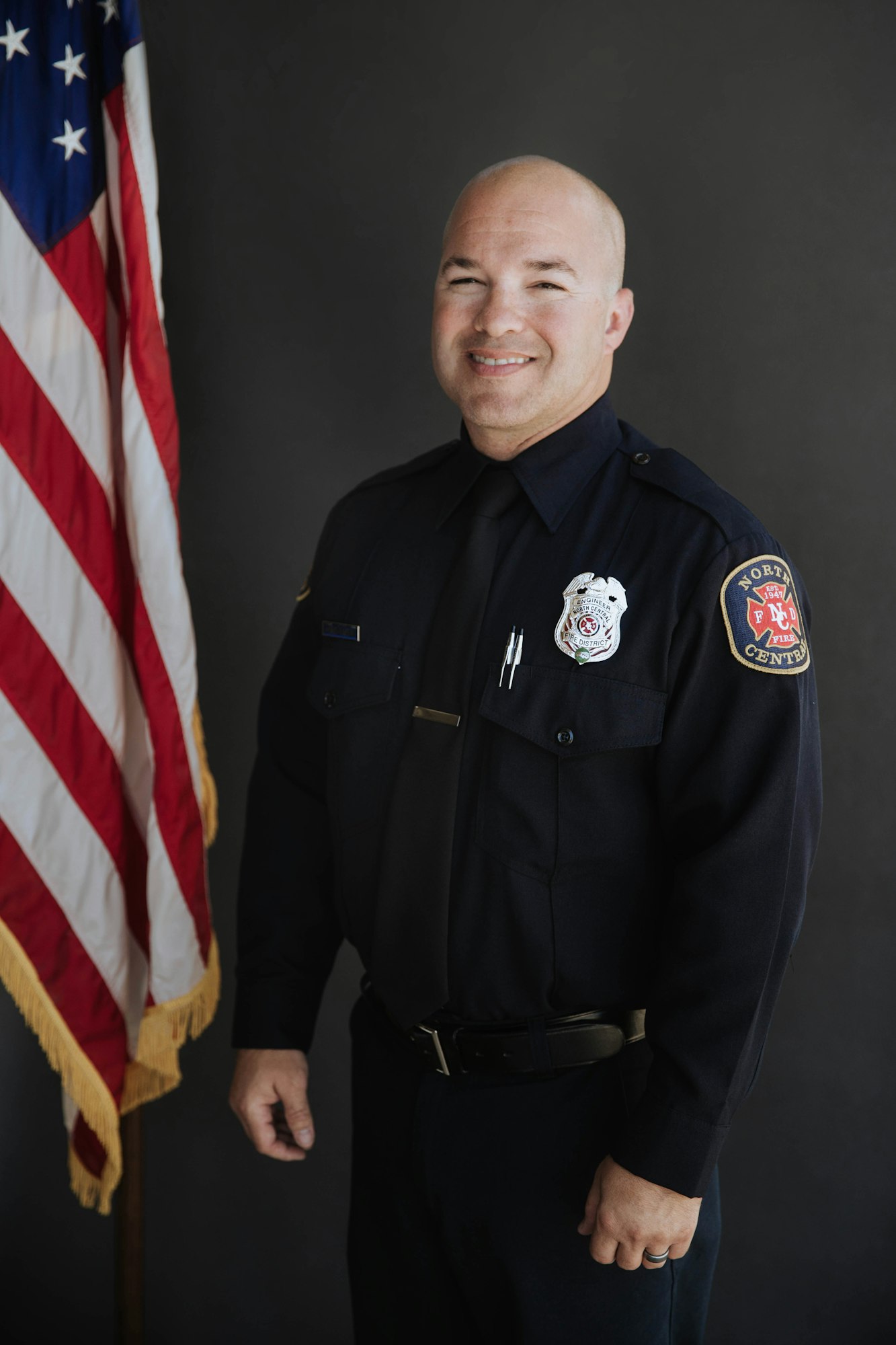 A firefighter stands smiling in uniform beside a U.S. flag.
