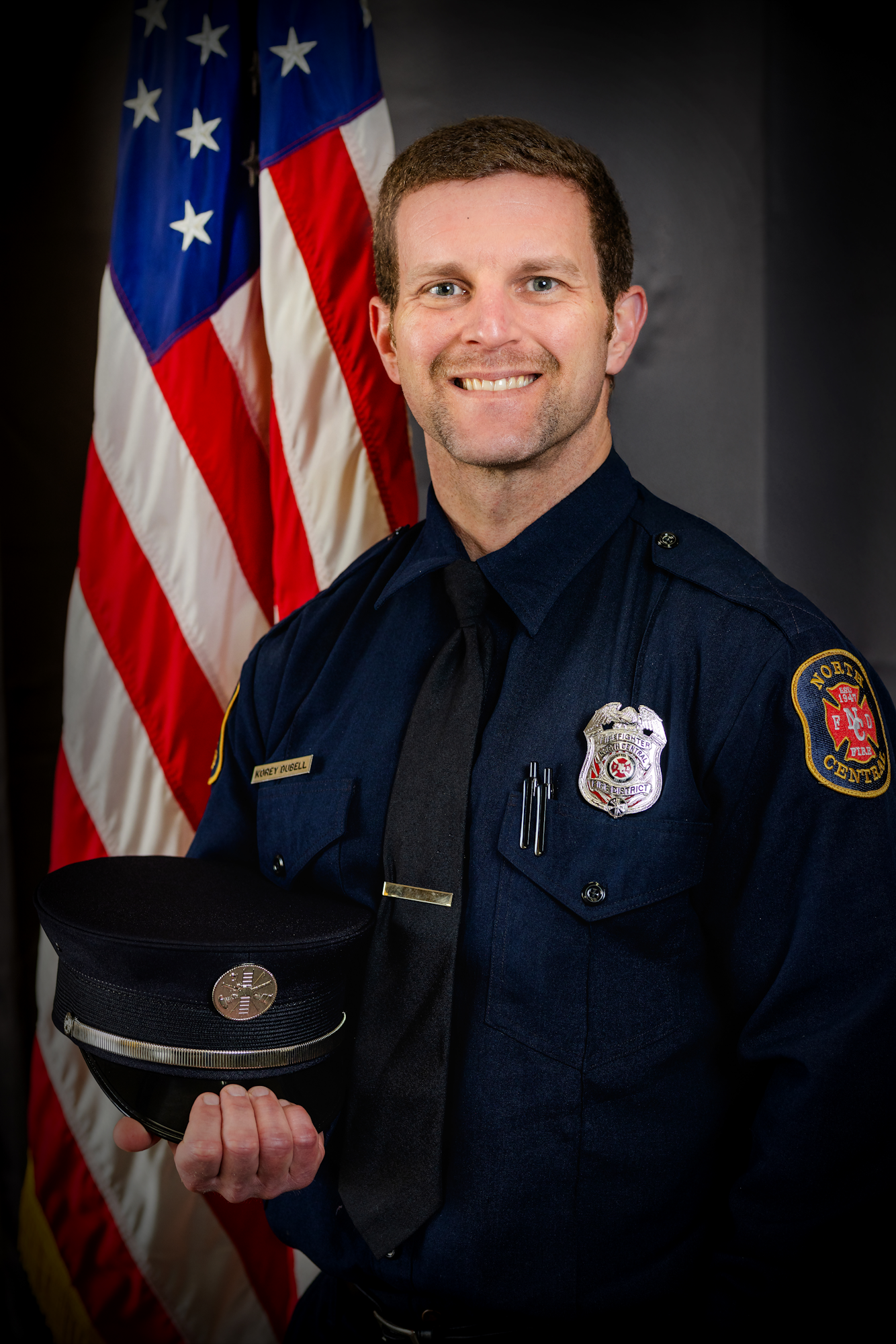 A smiling firefighter in a dark uniform stands in front of the American flag.