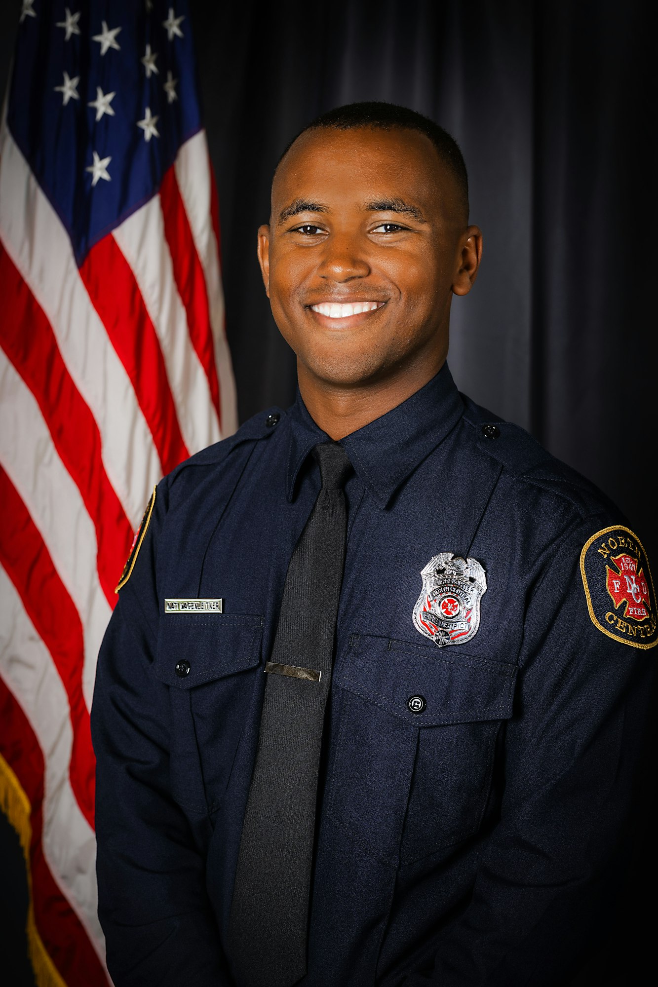 A smiling man in a firefighter uniform with an American flag in the background.