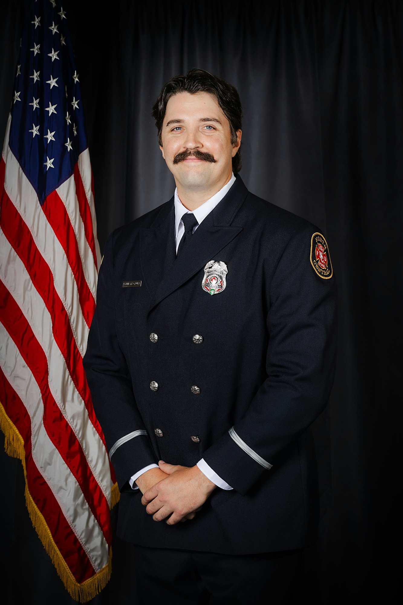 Fire Engineer Gauer in dress uniform stands before an American flag.