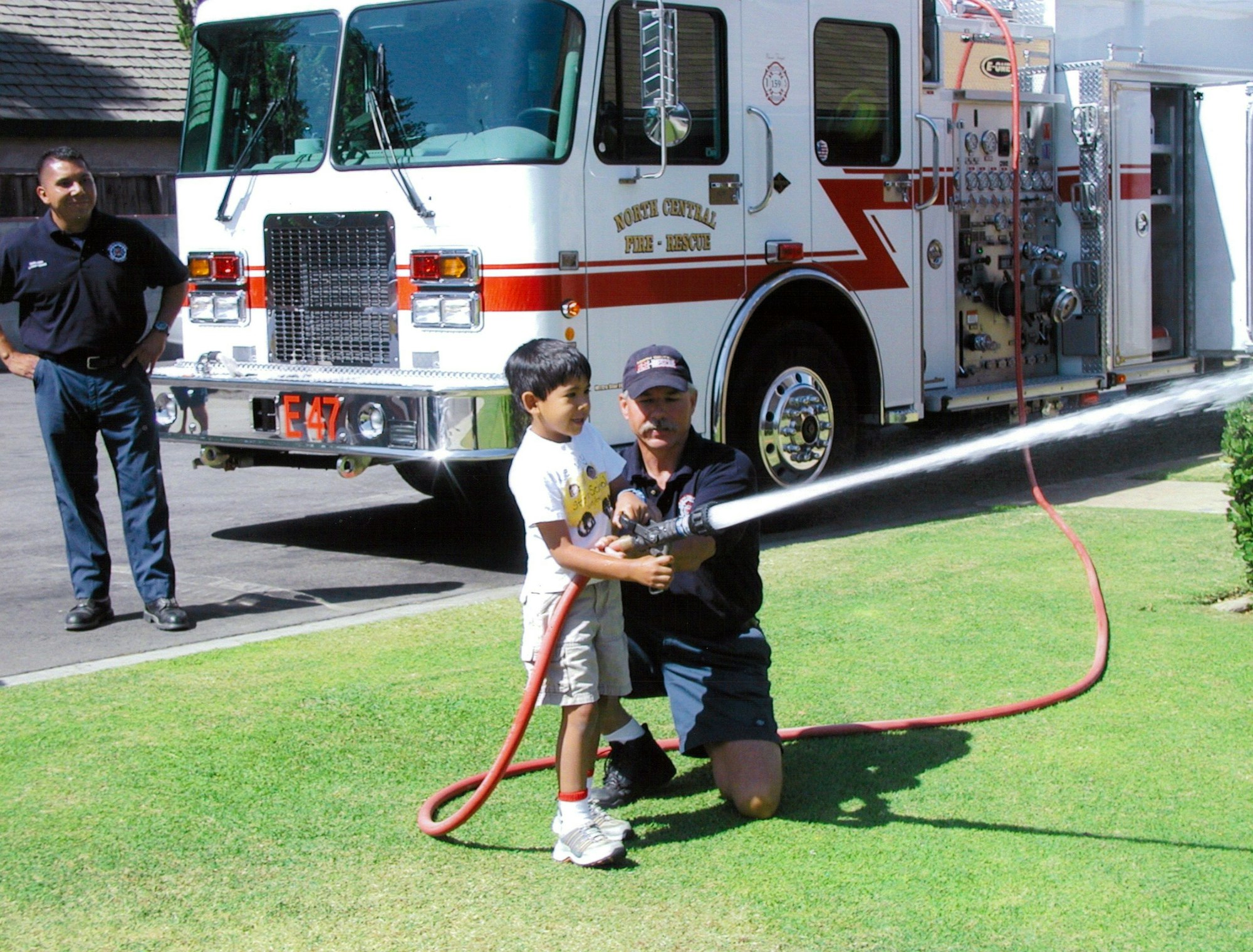 A child holds a fire hose with an adult firefighter, while another firefighter watches. A fire truck is parked nearby.