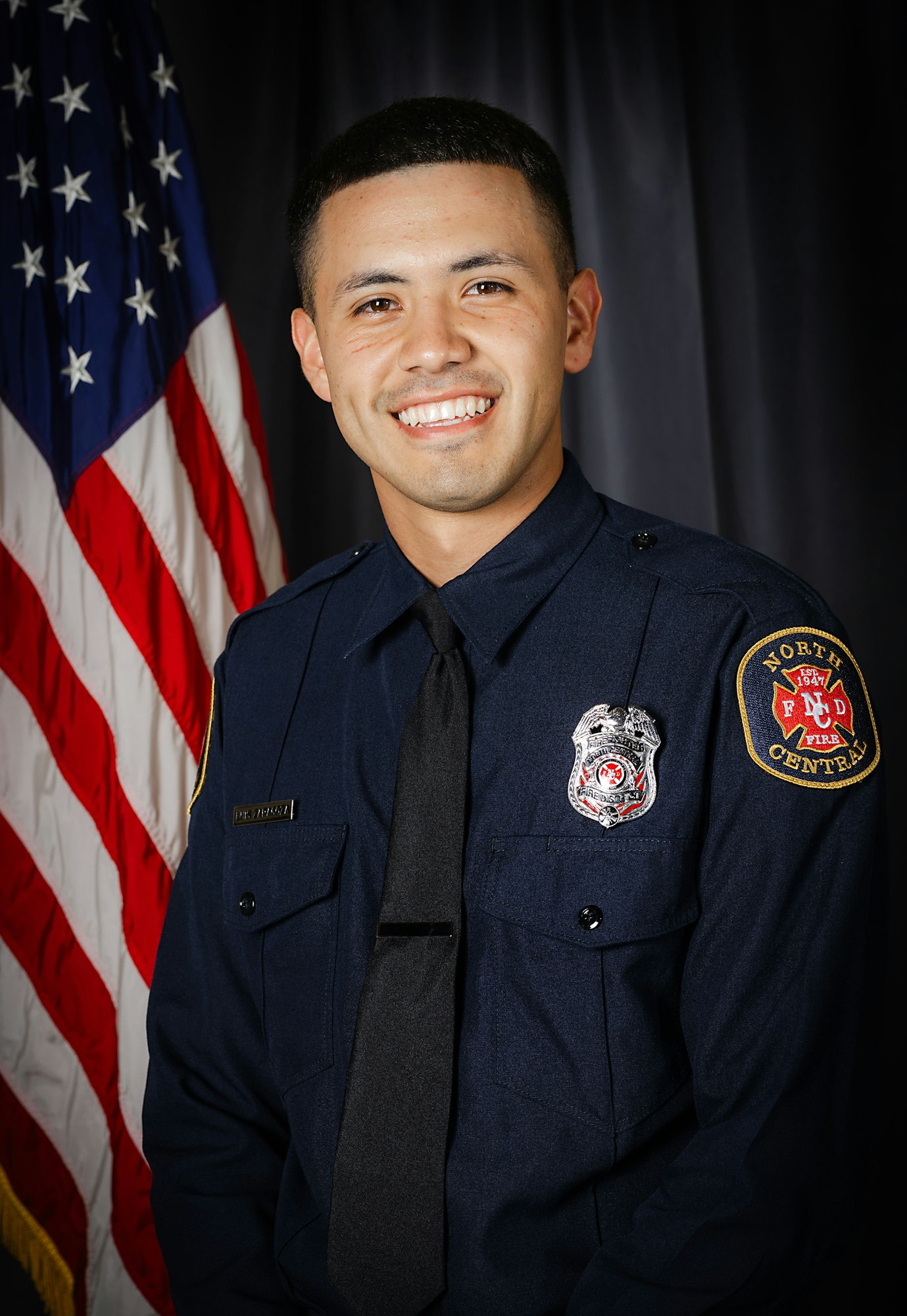 A smiling person in a firefighter's uniform with a badge, in front of an American flag.