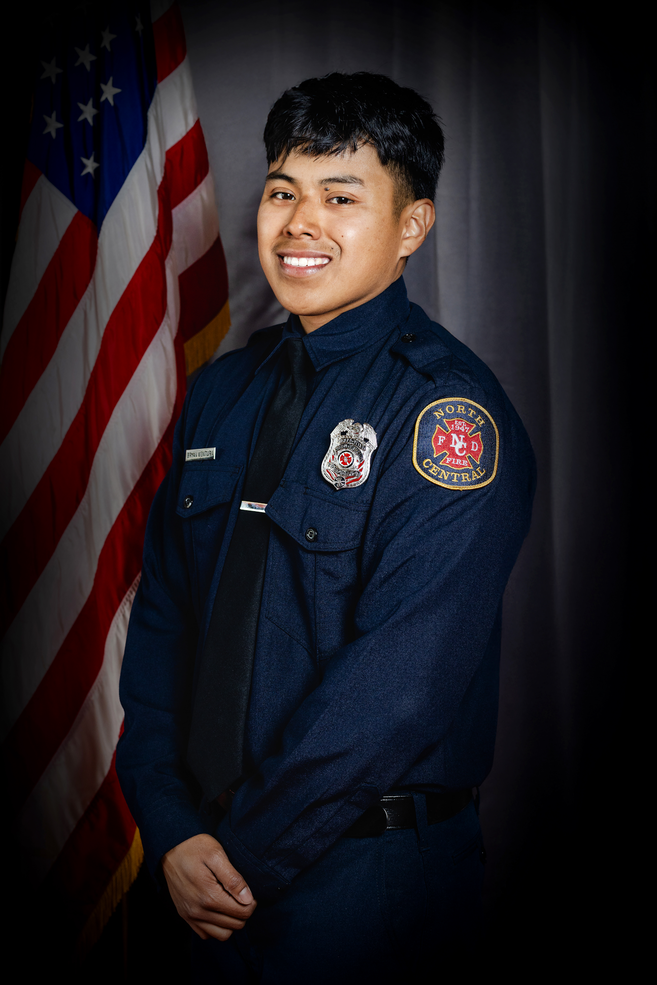 Bryan Ventura in a dark uniform stands beside an American flag, against a dark curtain background.