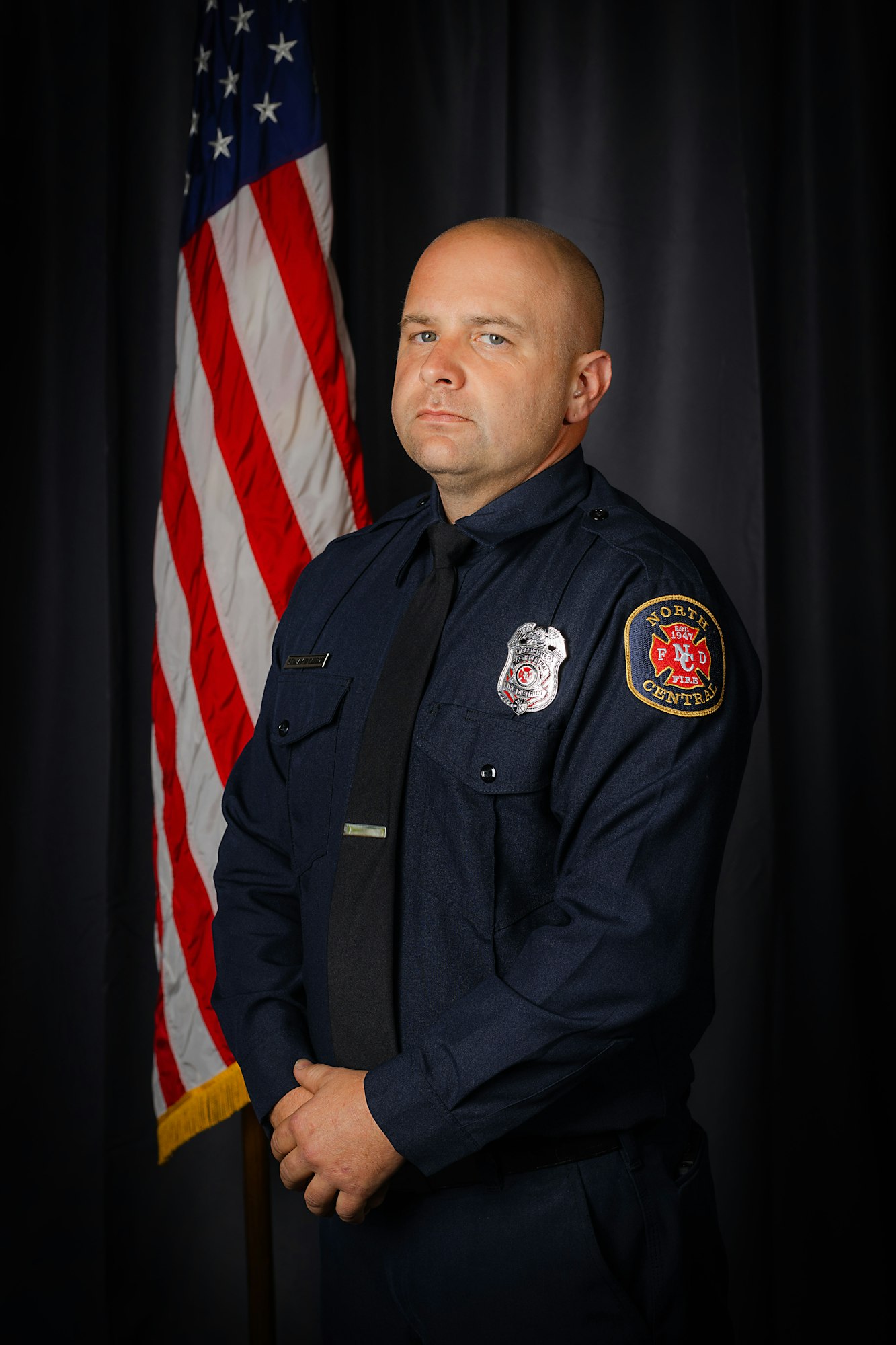 A firefighter in uniform stands in front of a U.S. flag, with a serious expression.