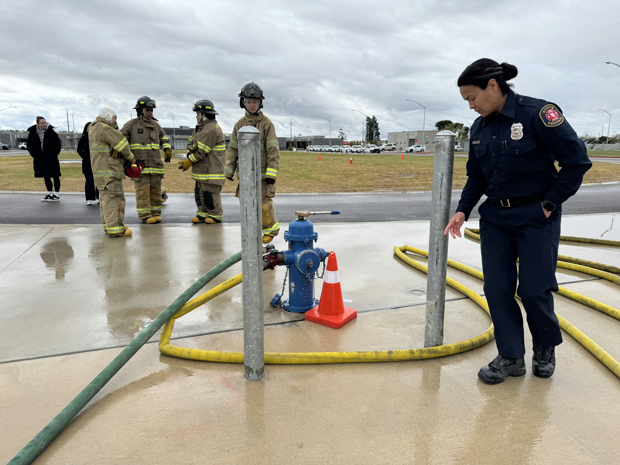 Firefighters and a fire hydrant with hoses, cones, and wet pavement, on a cloudy day.