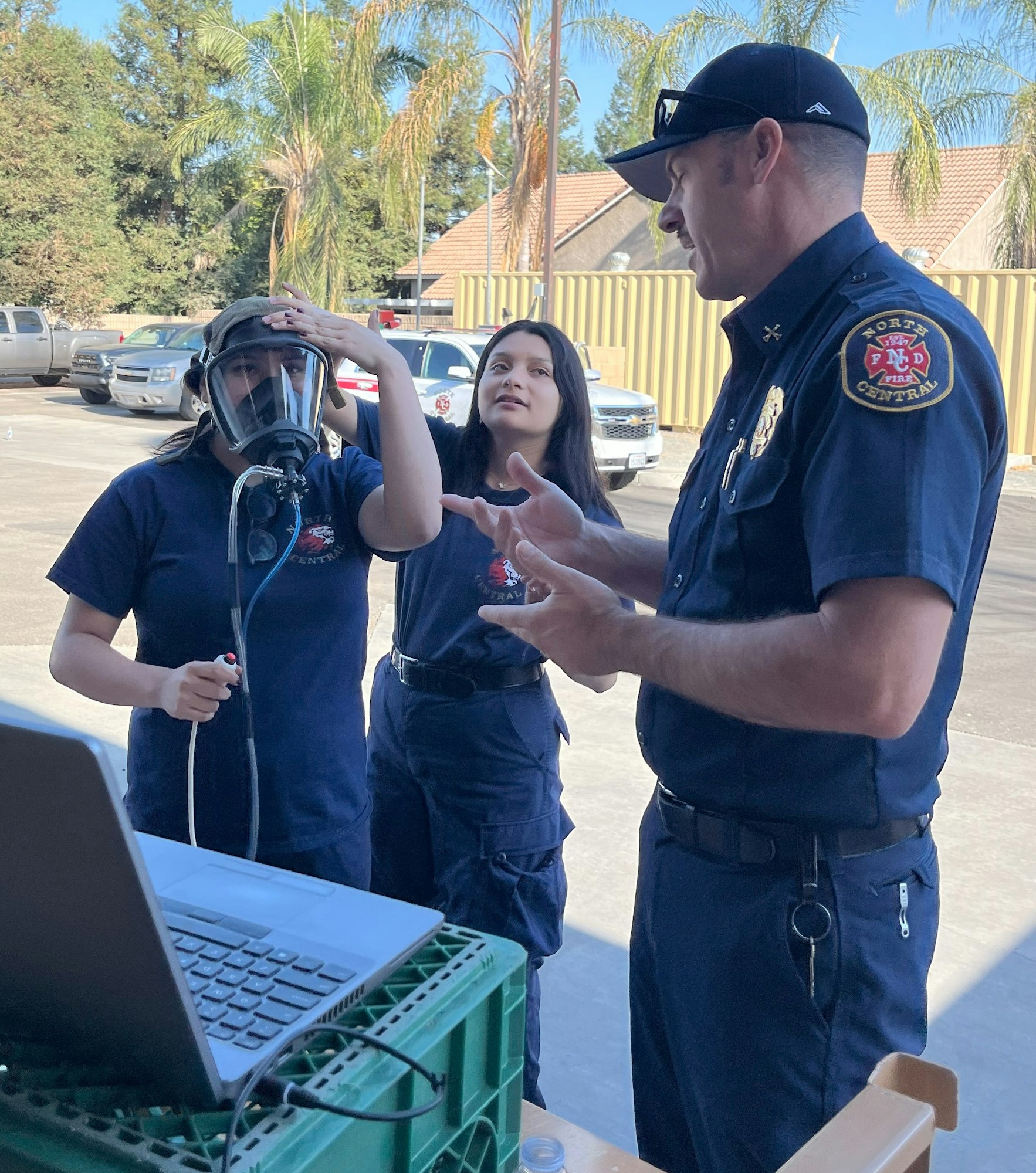Three individuals in uniform, one in a breathing mask, engaged in discussion outside near a laptop, with vehicles and trees in the background.
