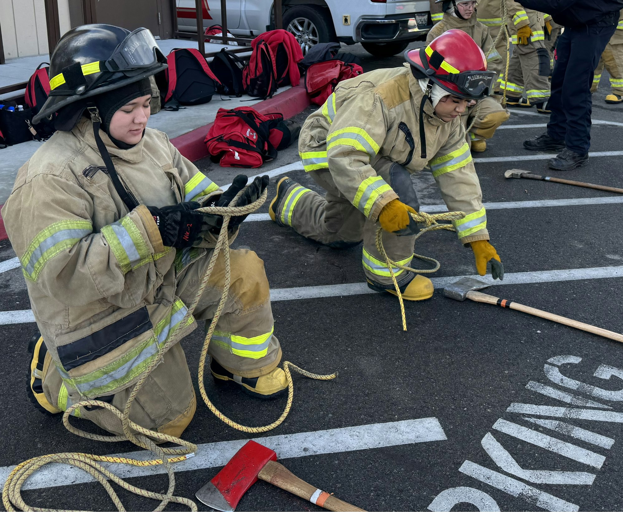 Firefighters kneeling, handling ropes with axes on the ground, in a practice setting.