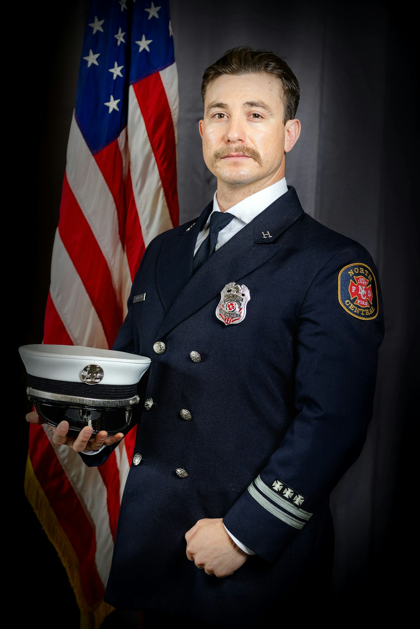 A firefighter in uniform holding a white hat stands in front of an American flag.